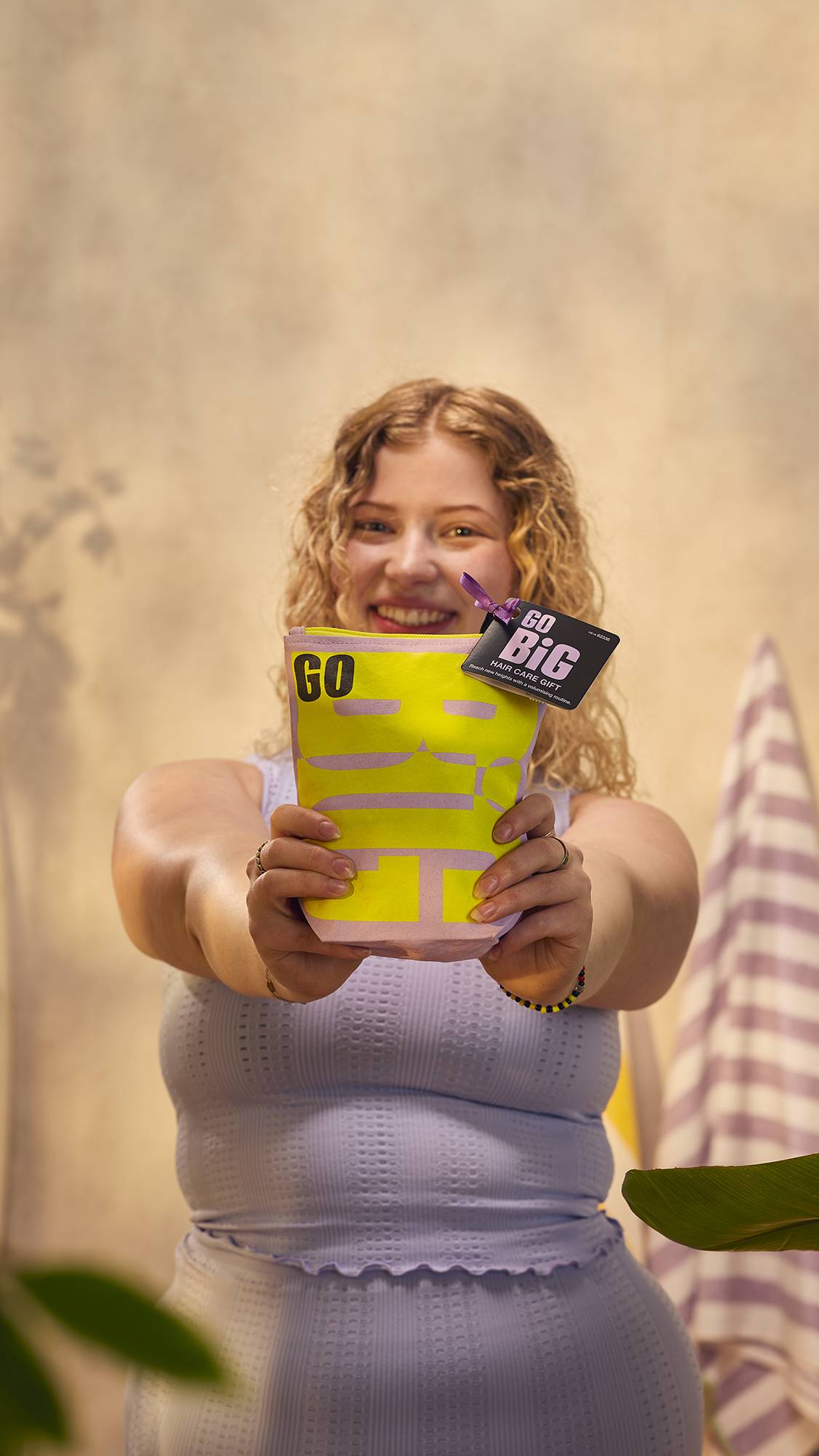 The image shows the model in a lavender-purple loungewear set, with curly blonde hair on a warm, earth-toned background holding the Go Big haircare gift bag up to the camera.