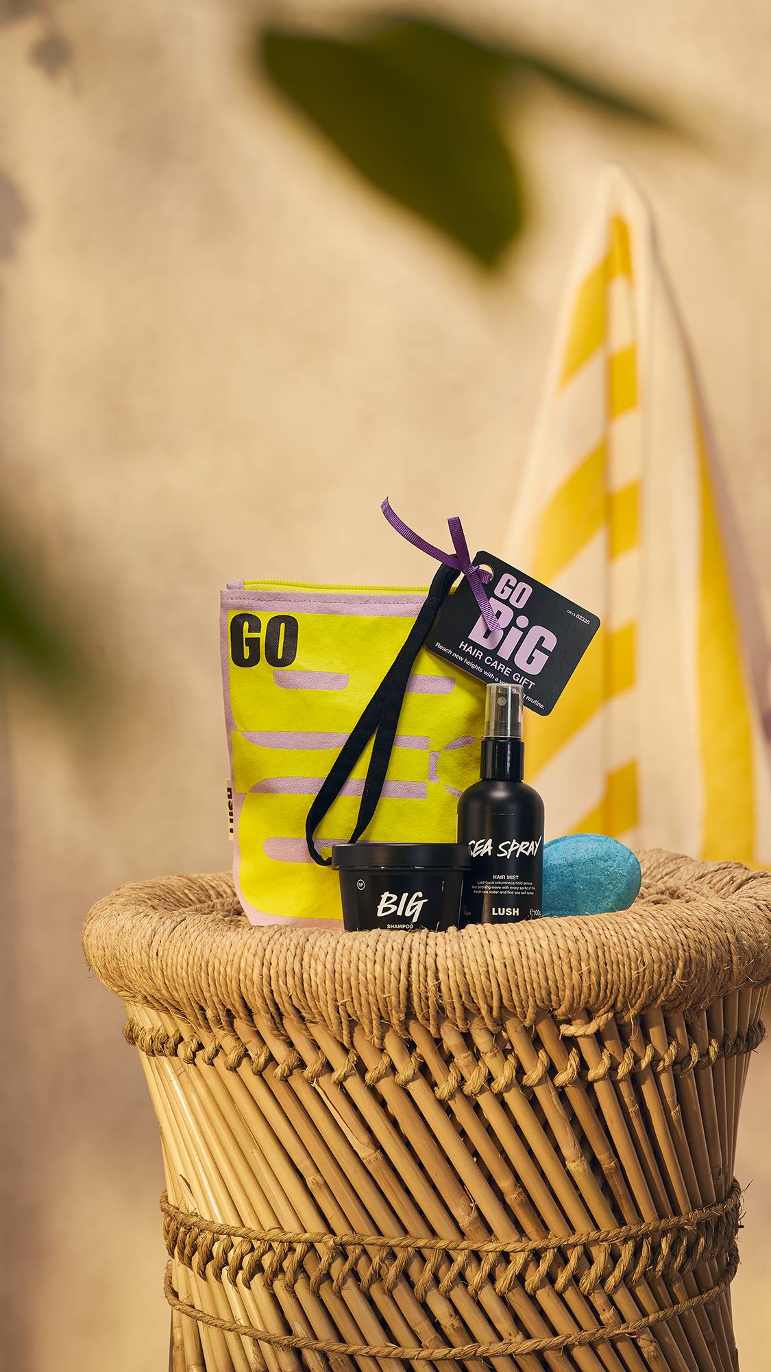 The image shows the Go Big pouch bag along with the three included haircare products sitting on top of a wicker hamper on a warm, earth-toned background. 