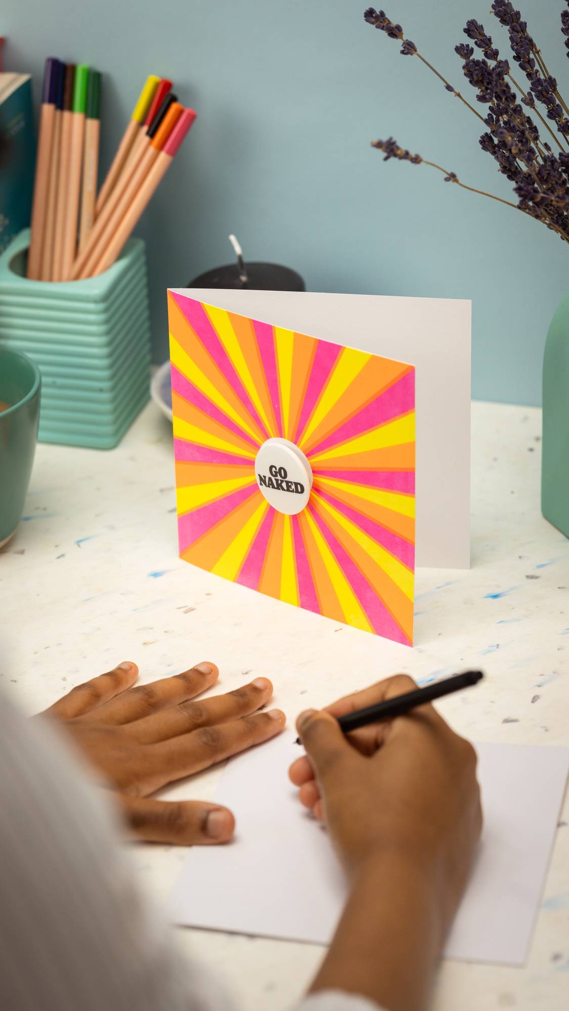 The colourful "Go Naked" card is stood up as the model writes the envelope on the table. The badge is still attached.
