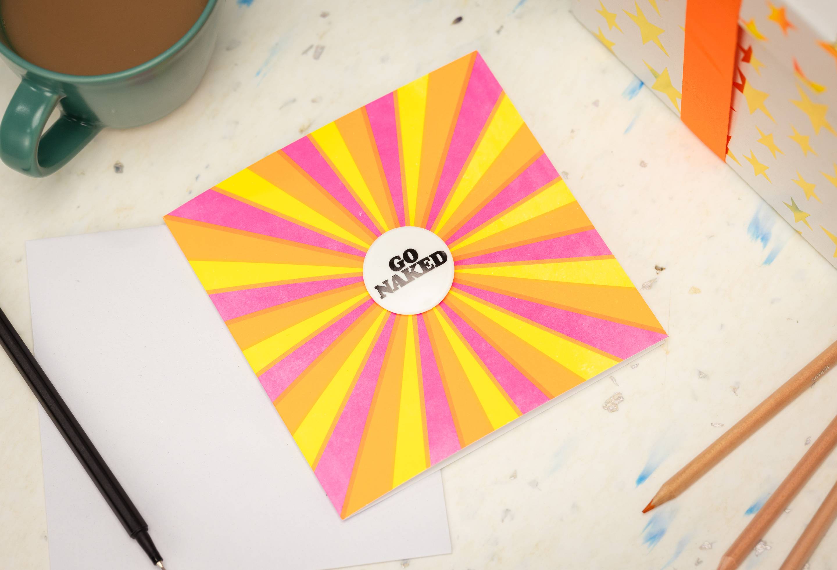 The vibrant "Go Naked" card and badge lay flat on a table surrounded by a wrapped gift, a pen and a mug of coffee.