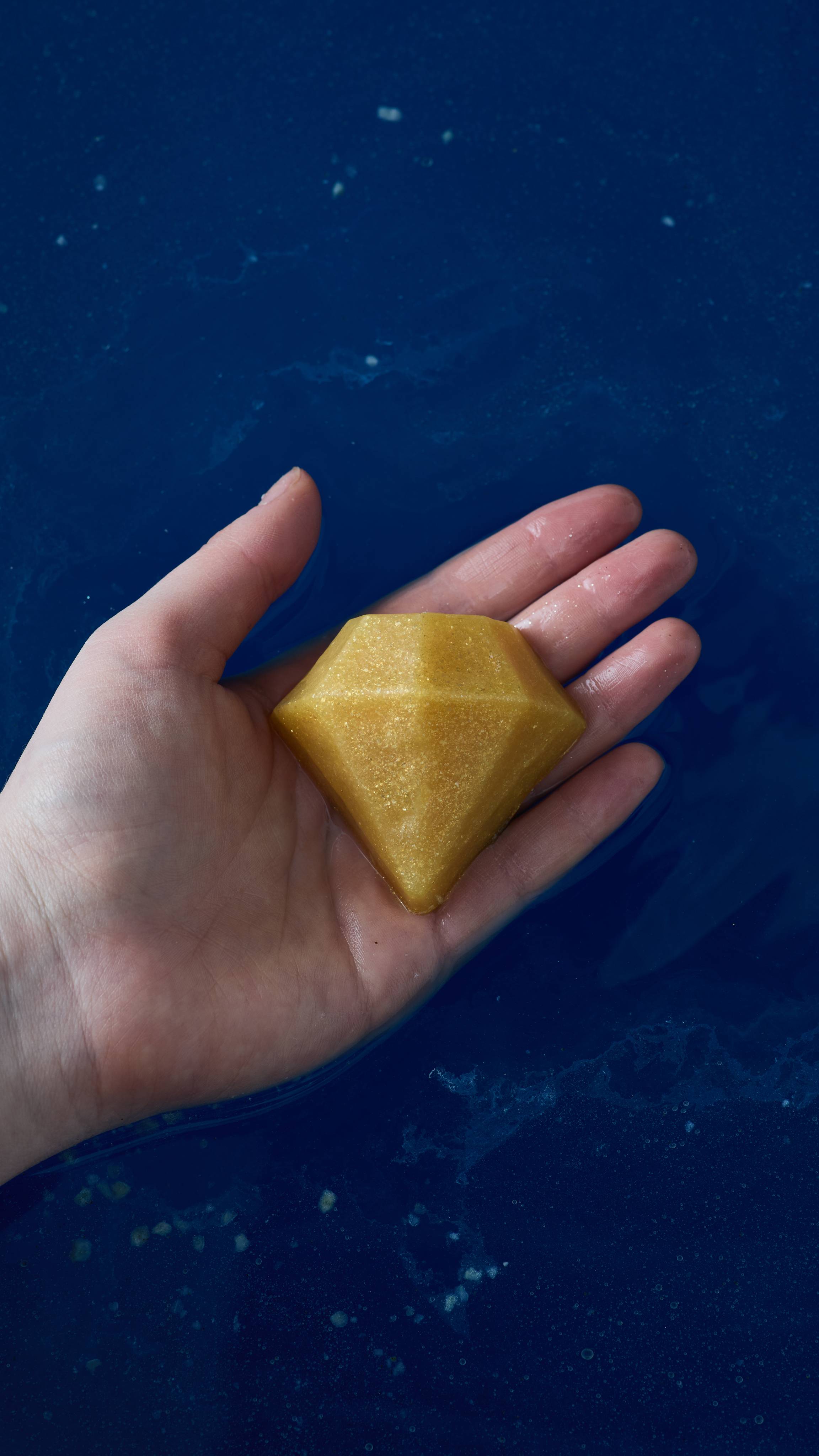 The image shows a close-up of the model's hand above deep, blue waters holding a golden, diamond-shaped soap. 