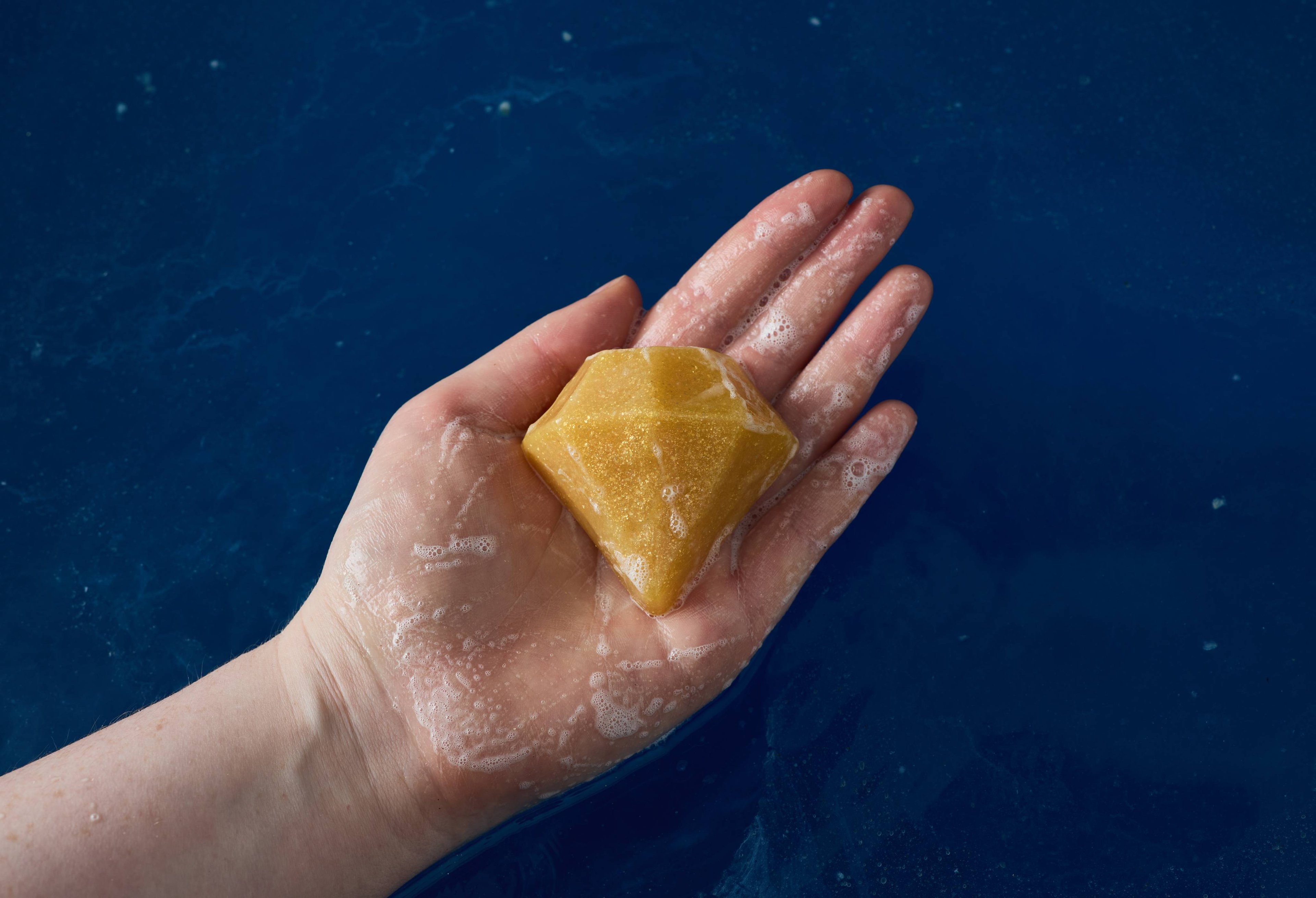 The image shows a close-up of the model's hand above deep, blue waters holding a shimmering, golden, diamond-shaped soap. 