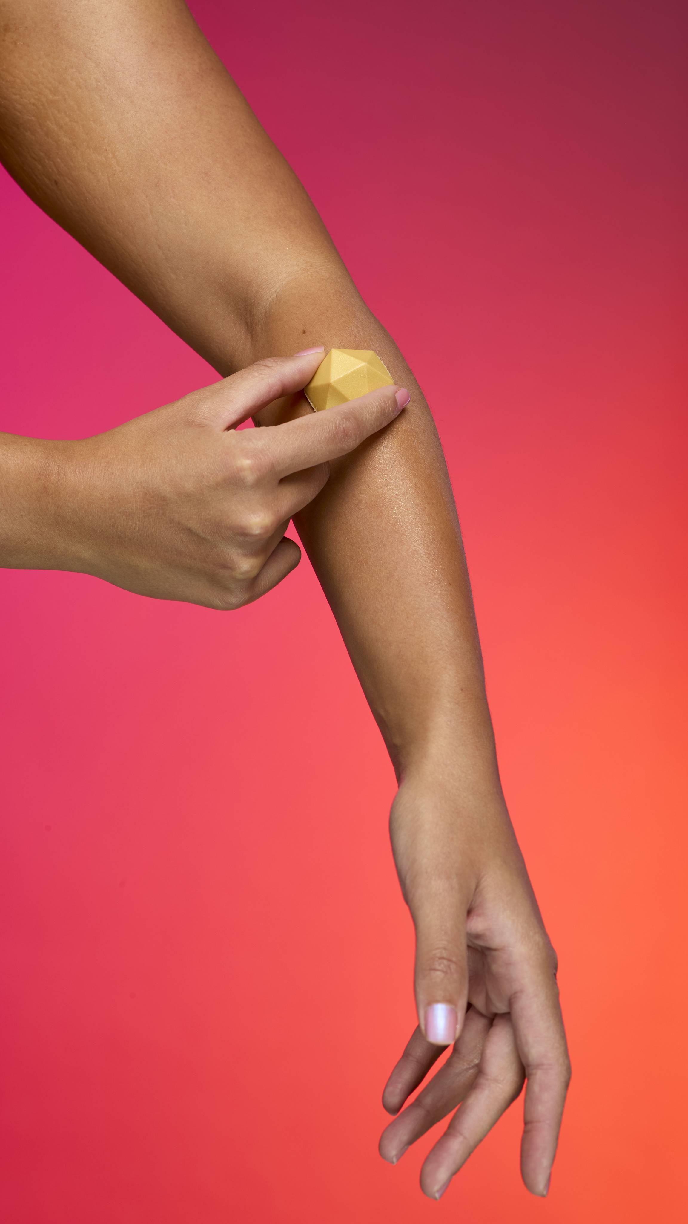 An arm with tan skin is shown as a hand holds and rubs the faceted, pyramid-shaped Gold Glitterbug body bar onto the forearm. A sheen of plastic free glitter is visible on the skin. The background is a vibrant gradient of magenta, pink, and orange.
