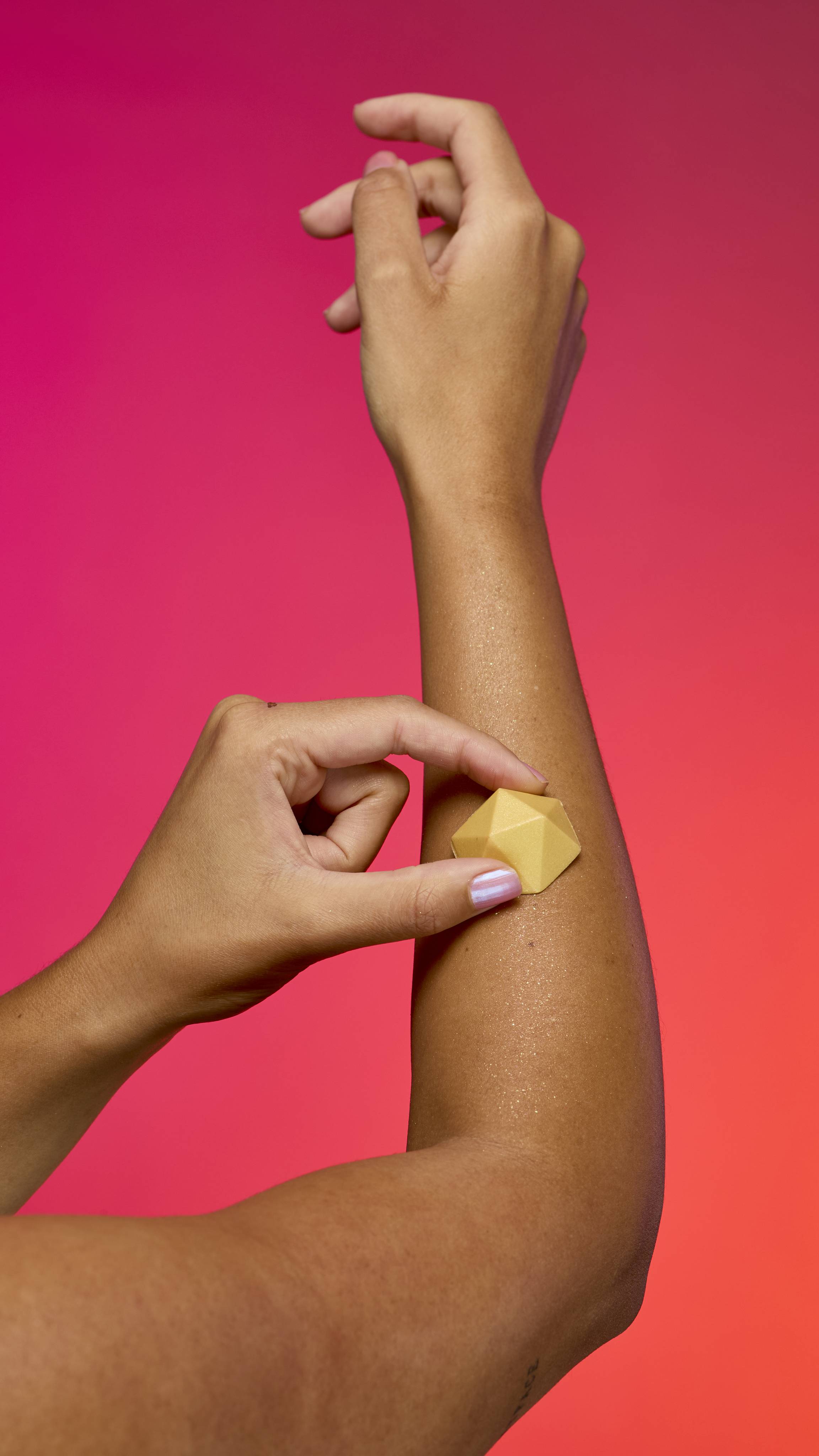 Two hands with tan skin hold and apply the faceted, pyramid-shaped Gold Glitterbug body bar to an arm. A visible sheen of gold plastic free glitter remains on the skin. The background is a vibrant gradient of magenta, pink, and orange.