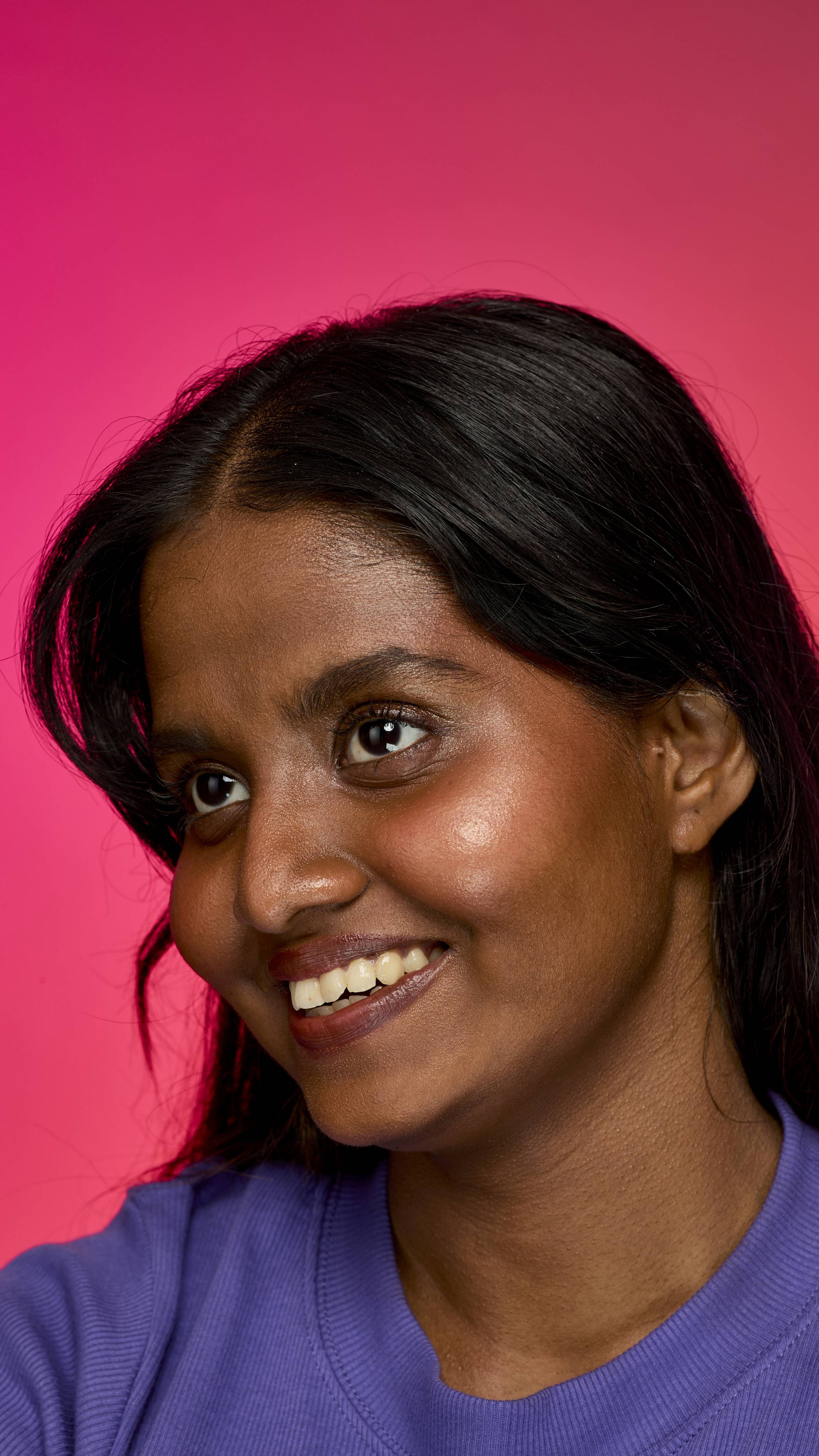 The image shows a dark haired model on a hot pink background. The model is wearing a purple sweatshirt as they smile showing the shimmery, golden glow on their cheekbones of the Gold Glow Stick highlighter.