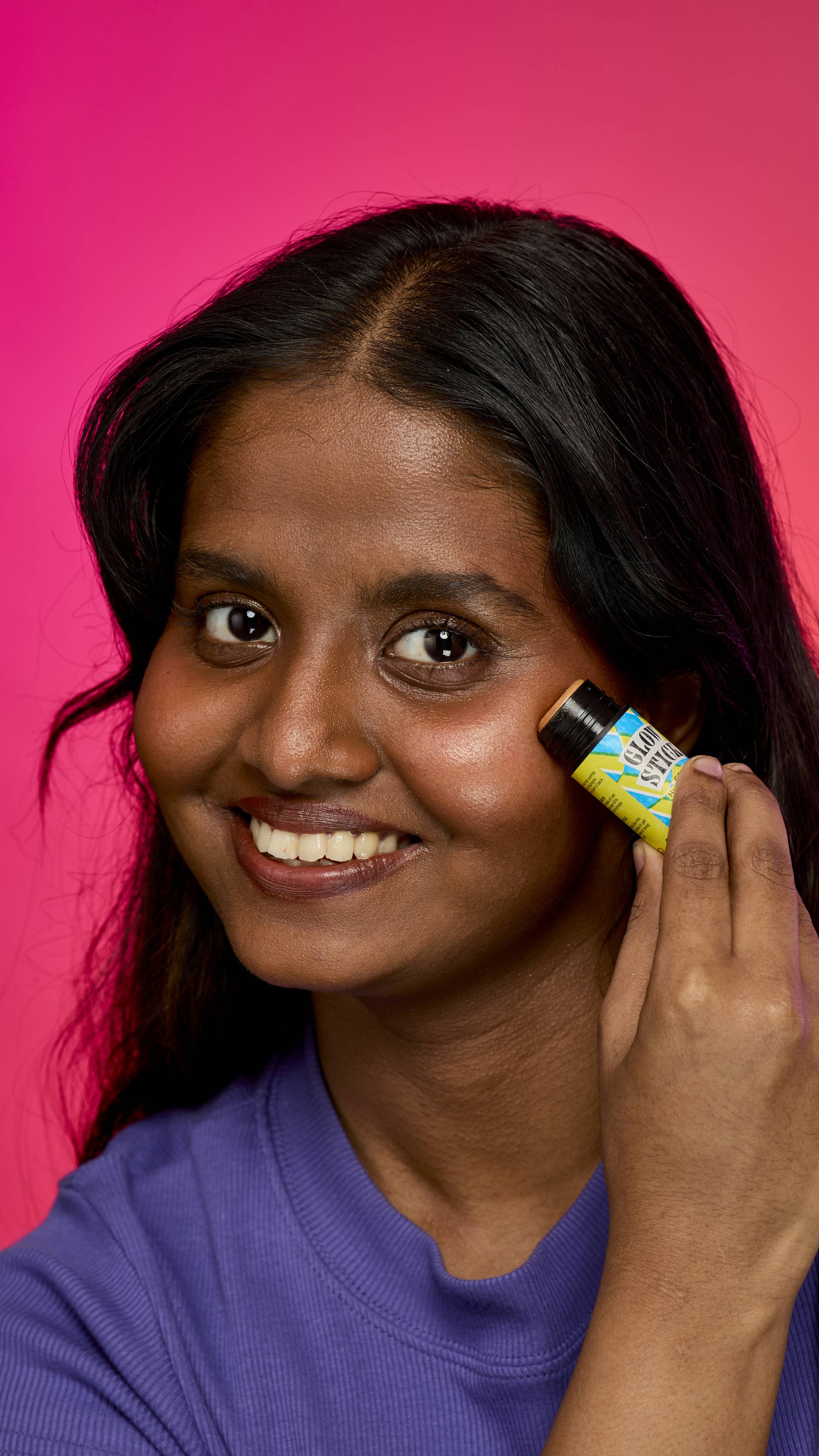 The image shows a dark haired model on a hot pink background. The model is wearing a purple sweatshirt and smiling as they apply the Gold Glow Stick highlighter directly to their cheeks. 