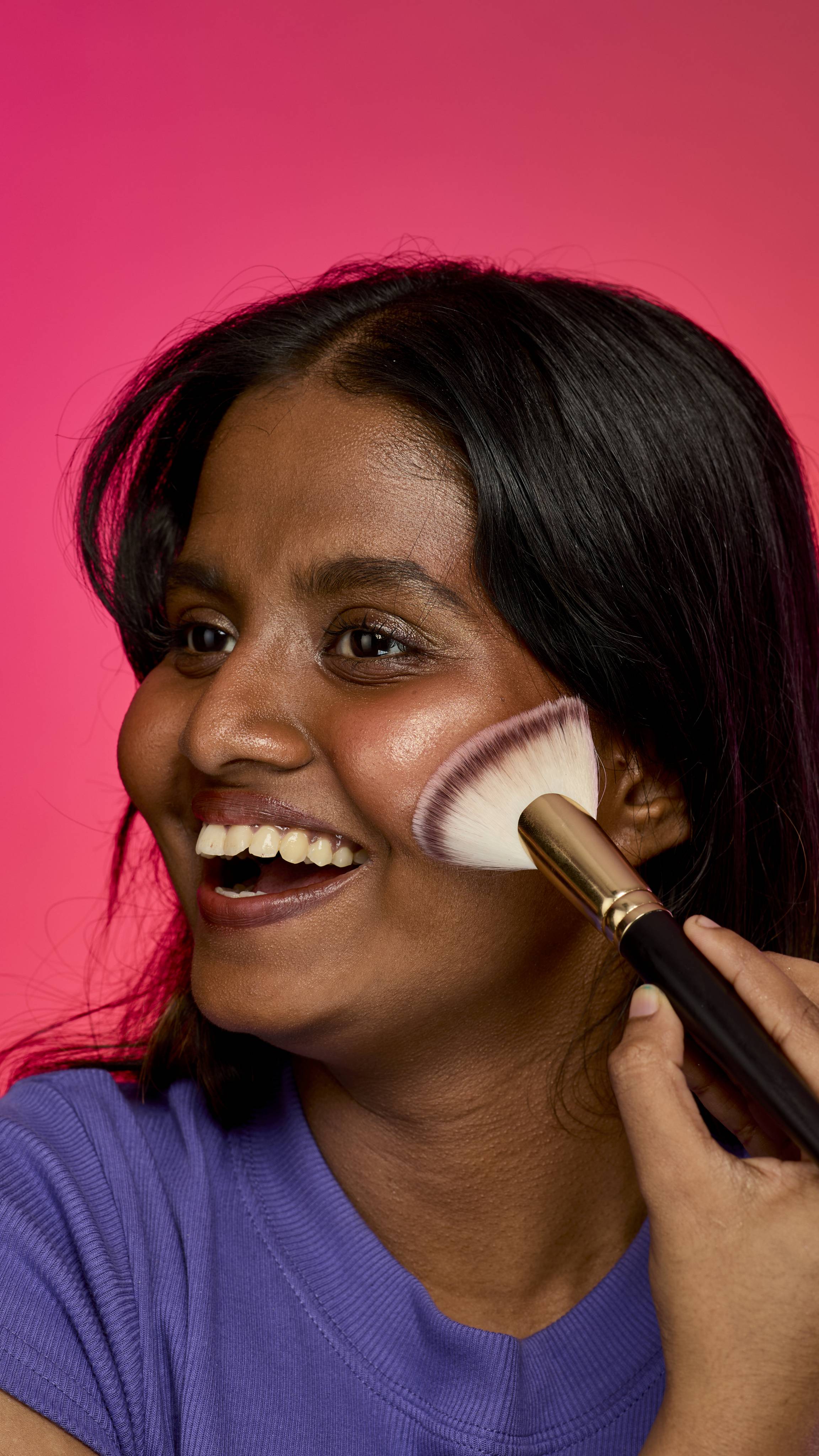 The image shows a dark haired model on a hot pink background. The model is wearing a purple sweatshirt and smiling as they blend the Gold Glow Stick highlighter on their cheekbones with a fanned brush.
