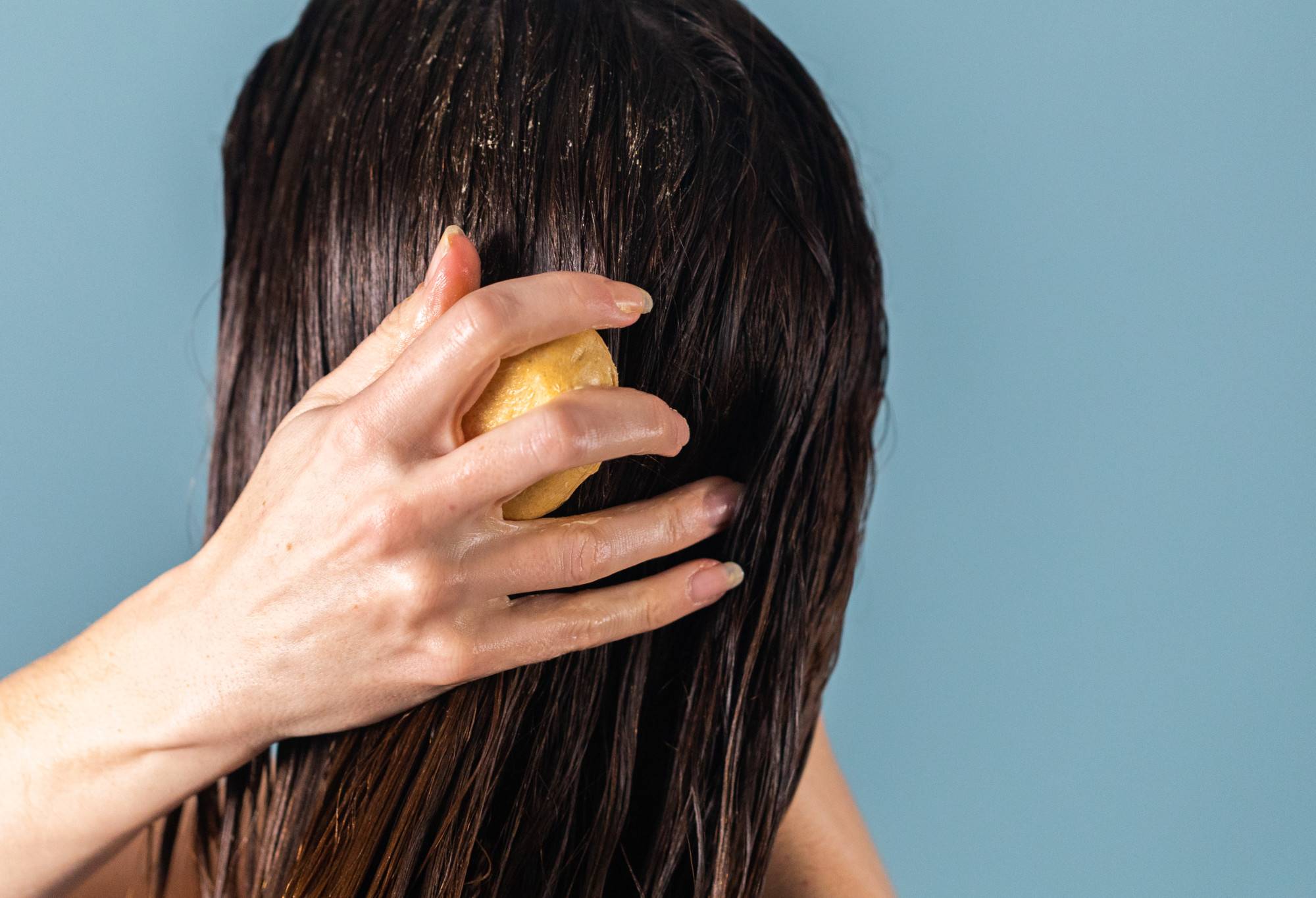 A person with long, brown hair rubs The Golden Cap, a yellow, oval shaped solid conditioner bar, through their hair.