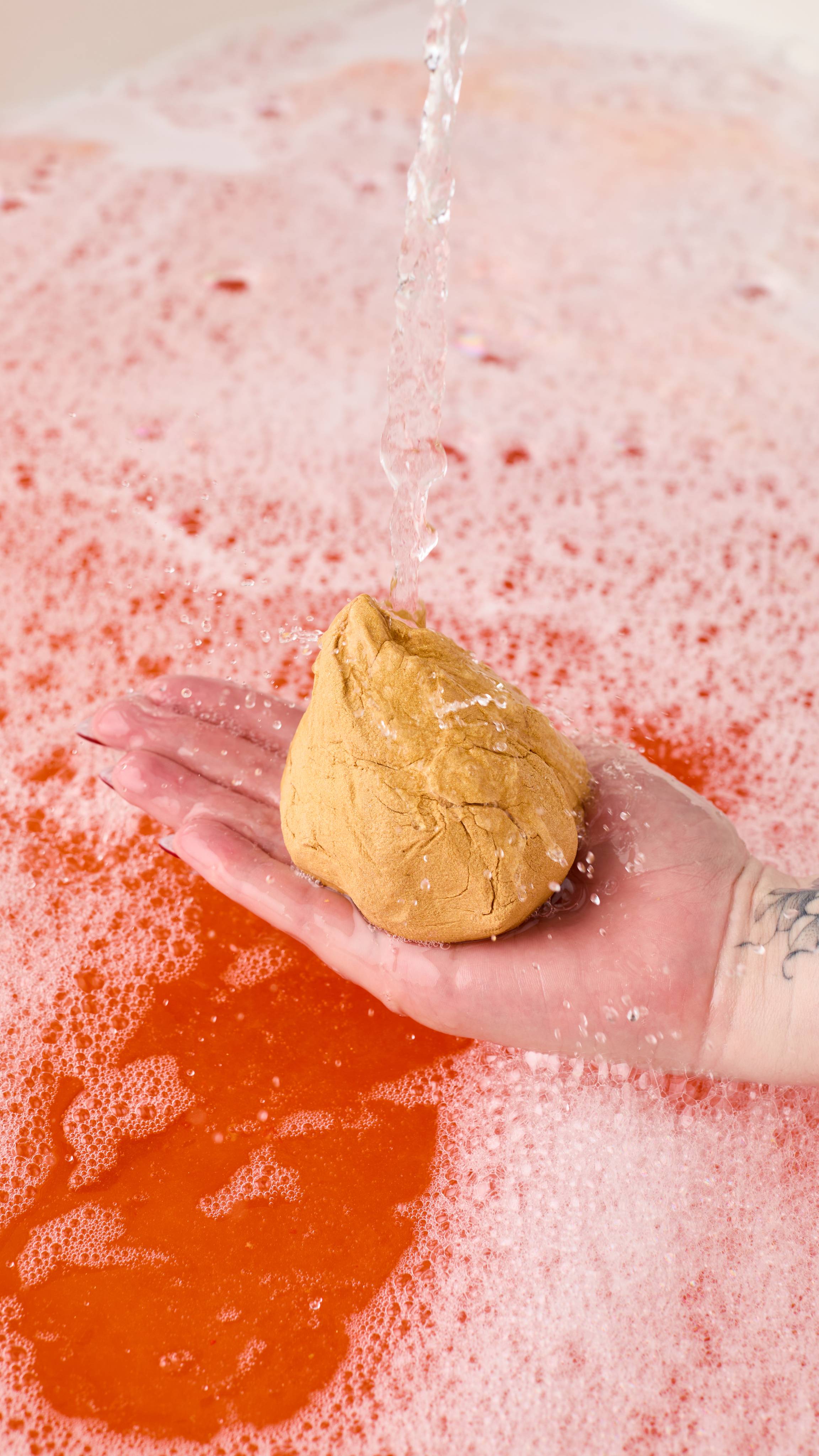 The image shows a light-toned hand holding a golden, sack-shaped Golden Wonder bubble bar under a strong stream of water. Red-orange bath water is visible beneath the bubbles below.