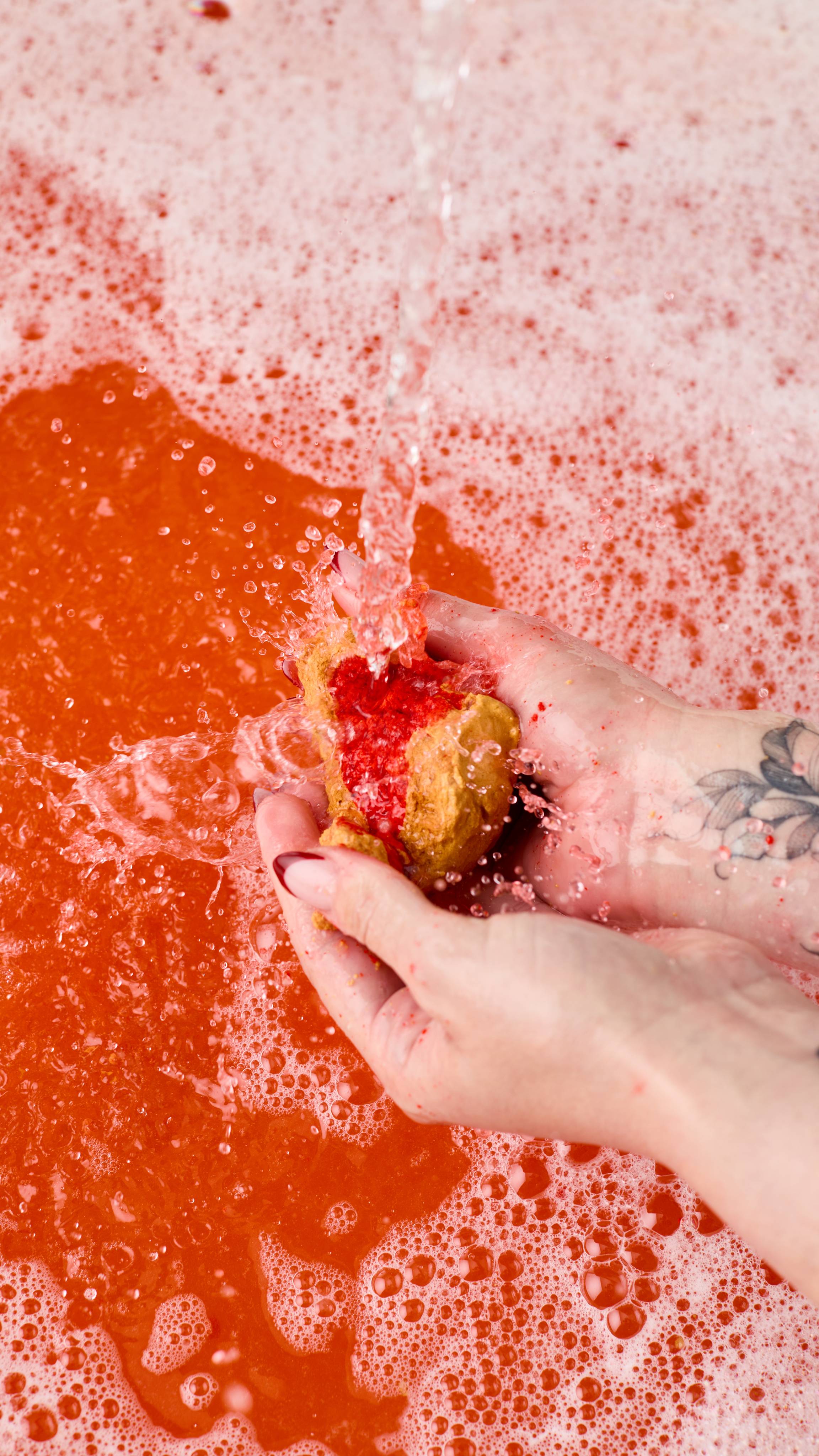The image shows two hands with long nails vigorously crumbling the Golden Wonder bubble bar. A bright red core is revealed as a strong stream of water creates abundant foam and splashes across the red bathwater.