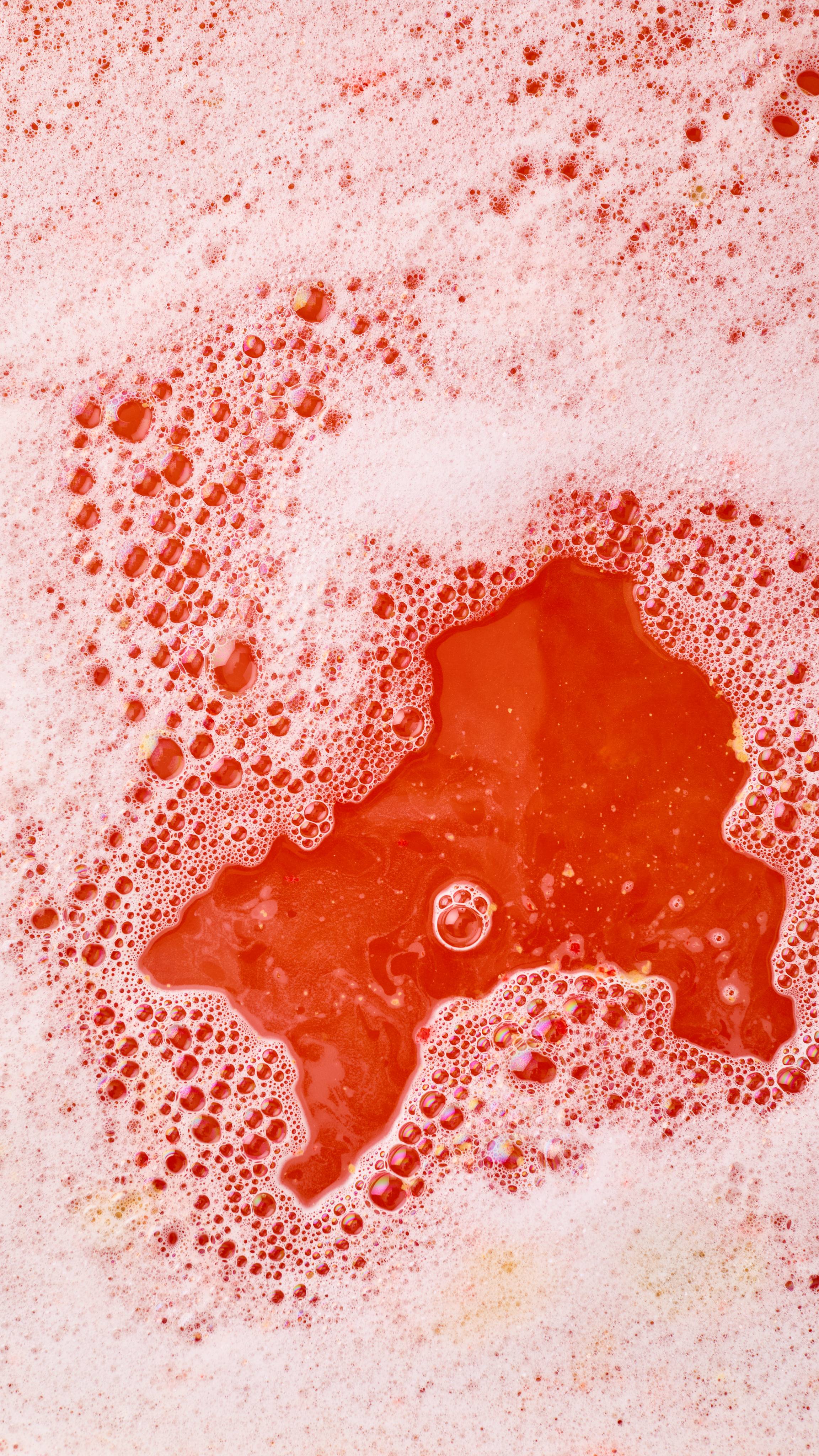 A close-up, aerial image of the bath water after using the Lush, Golden Wonder bubble bar. There is a blanket of thick, fat bubbles with vibrant red bath water visible through a large gap.