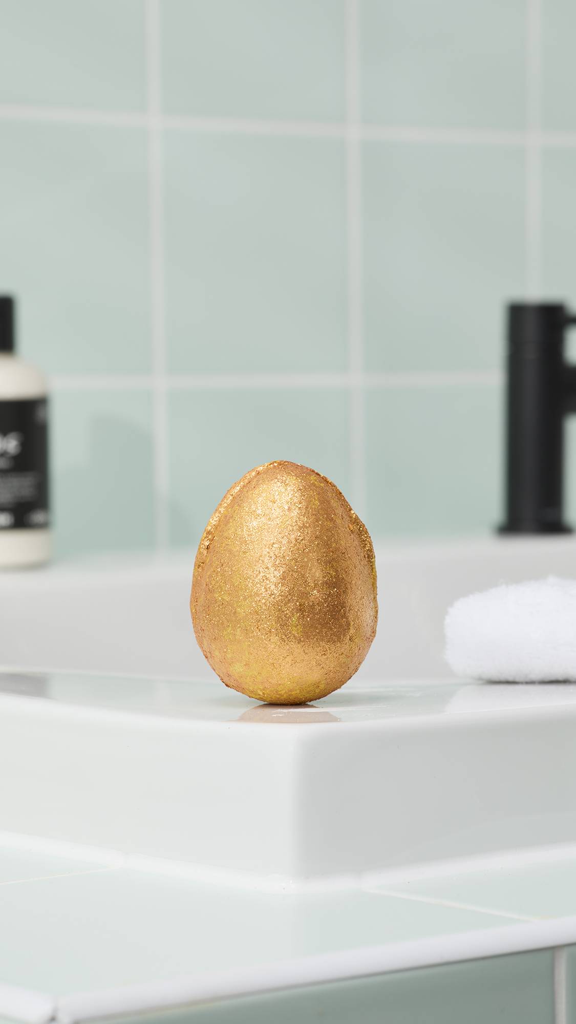 An oval, textured Golden Egg bath bomb coated in a vibrant, metallic gold shimmer sits on the edge of a white bathtub. The background features soft mint green tiles and a black faucet. All glitter used is plastic-free.