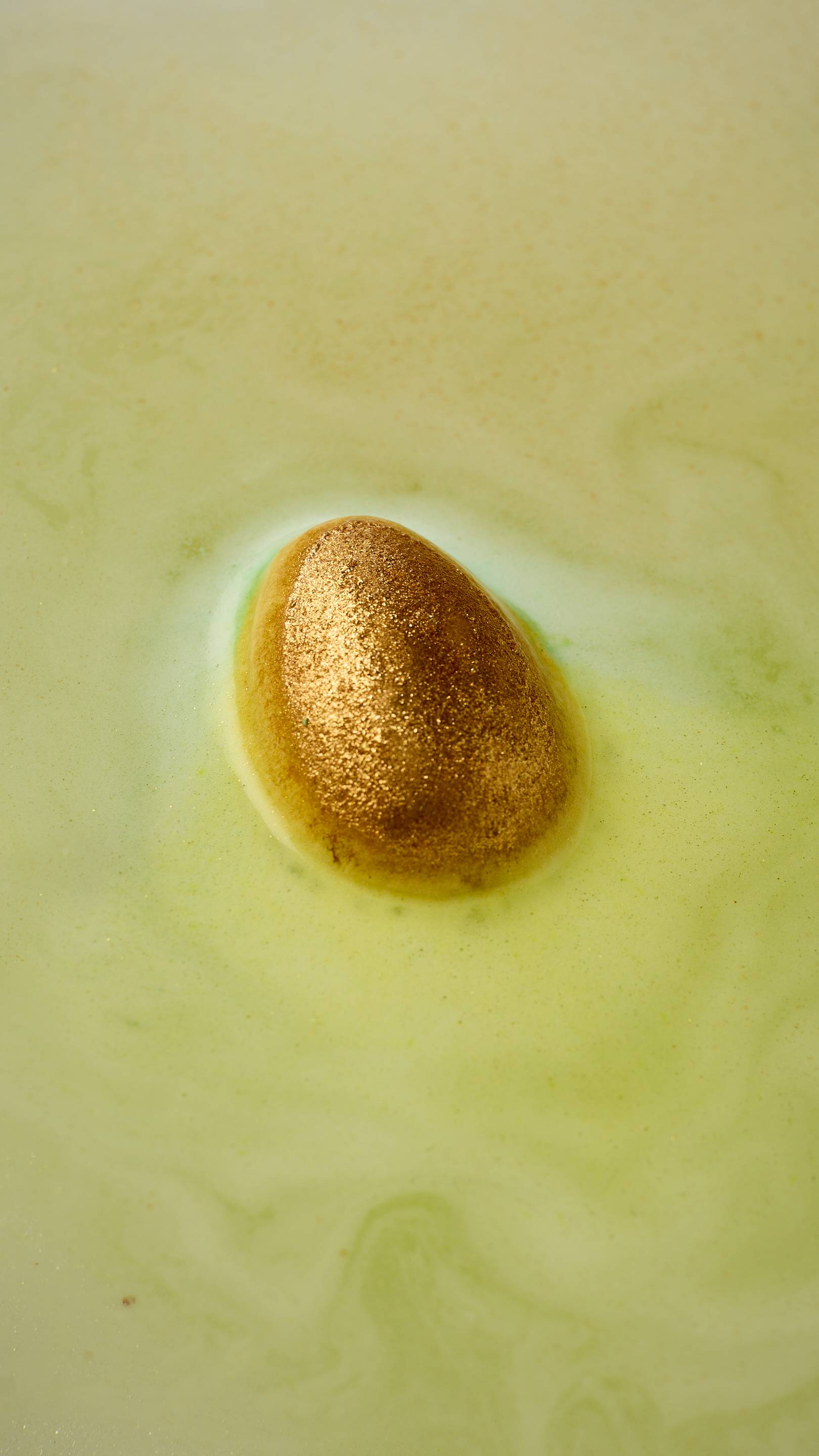 An aerial view image of the "Golden Egg' Easter-themed bath bomb from Lush. The bath bomb is a perfect egg shape, covered in thick, sparkling, gold lustre. The product begins to dissolve, creating soft, yellow bathwater. 