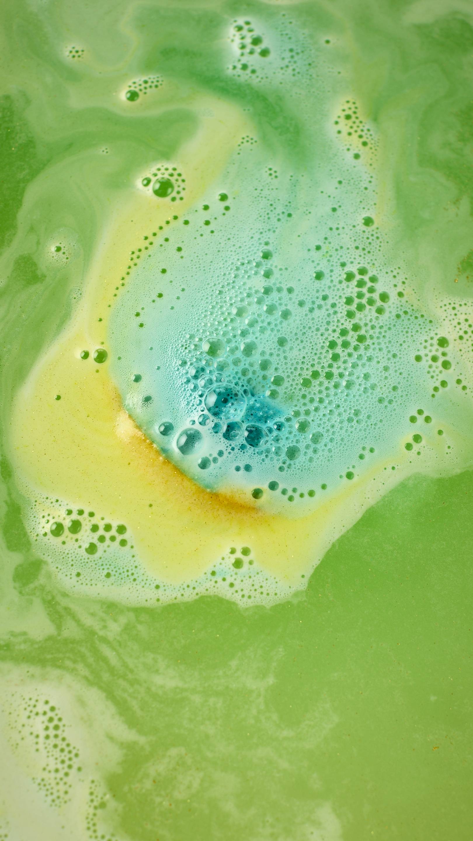 The Golden Egg bath bomb dissolves in a pool of vibrant green water, releasing a core of bright turquoise foam. Shimmering, plastic-free gold glitter sparkles throughout the swirling bubbles and neon-colored ripples.