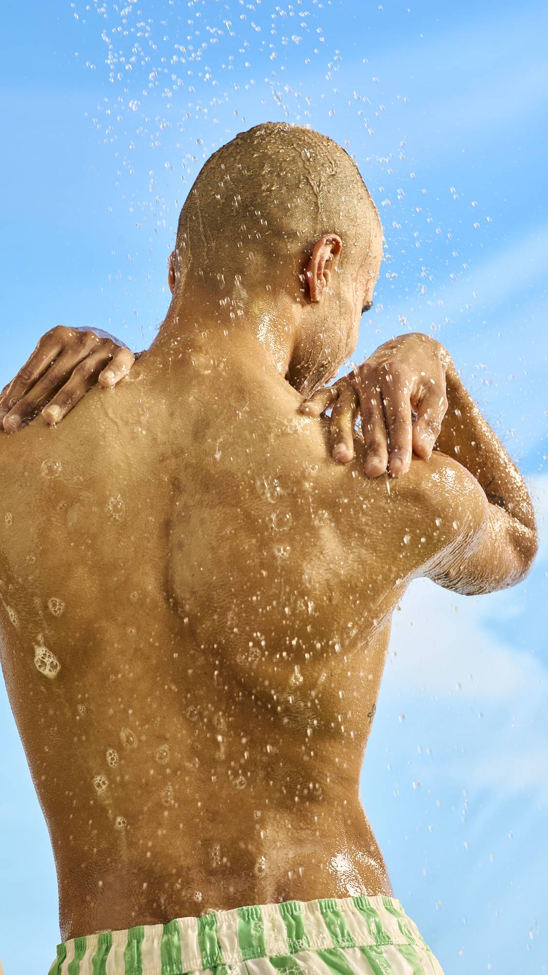 The model is standing under an outdoor shower under a bright blue sky with their back to the camera as soapy suds of Golden Egg shower gel trail down their back. 