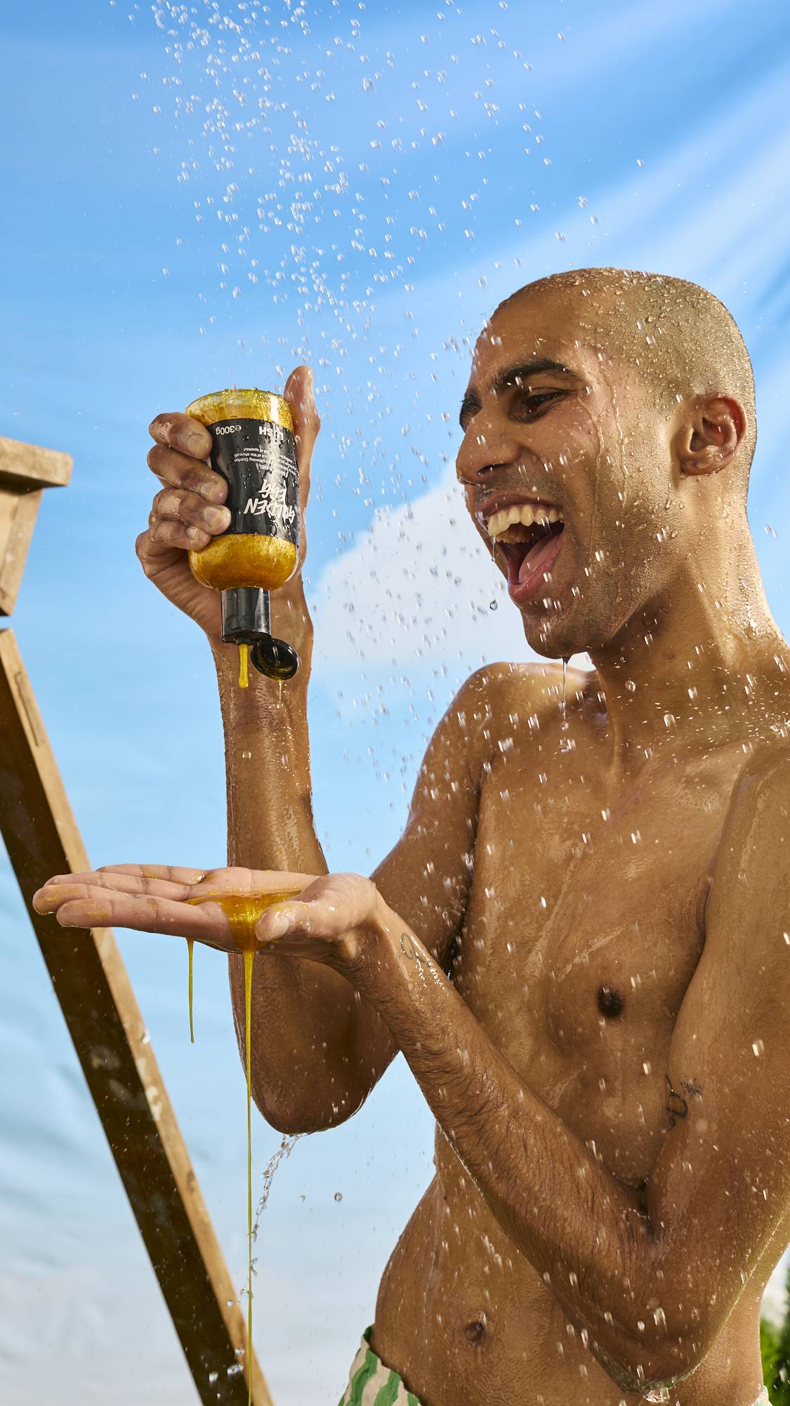 The model is standing under an outdoor shower under a bright blue sky as they are generously squeezing the golden, glittery shower gel into their palm. 