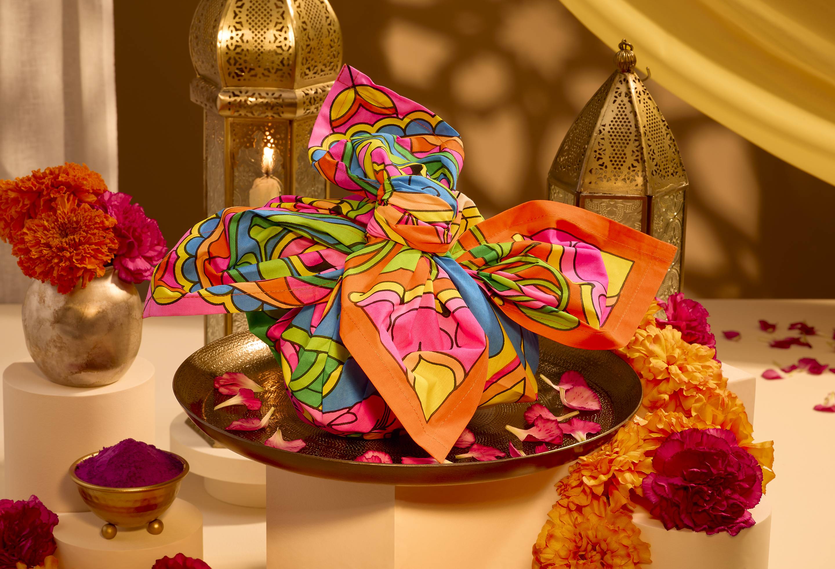 A gift wrapped in a vibrant, psychedelic pink, blue, yellow, and green "Golden Glow" Lush Knot Wrap sits on a bronze plate, framed by marigolds, scattered petals, brass lanterns, and purple pigment in a warm Diwali setting.