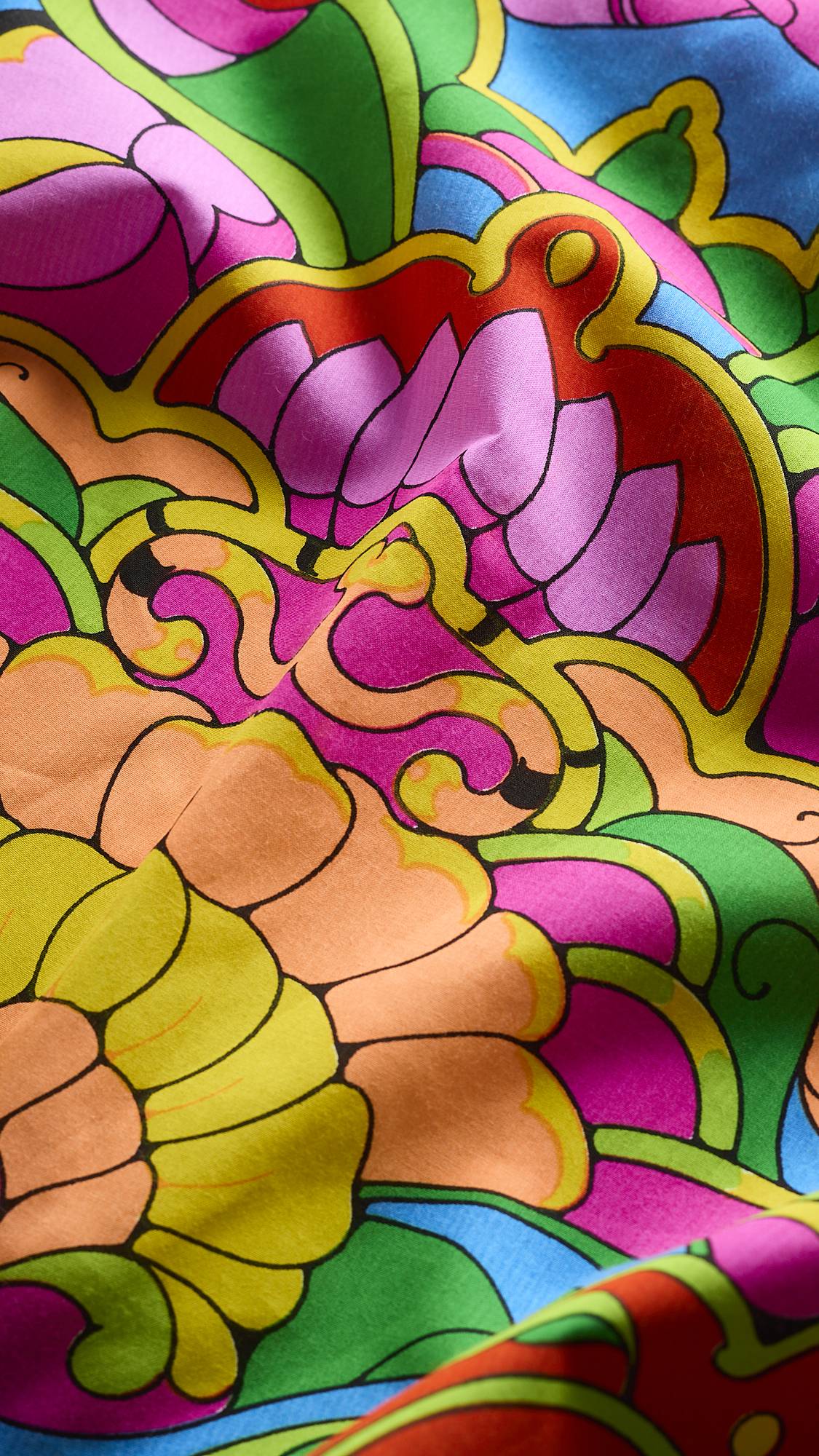 A close-up shot of soft, folded fabric from a Lush Knot Wrap, featuring vibrant stained-glass-style illustrations of lotus petals and decorative swirls in shades of pink, yellow, green, and orange.
