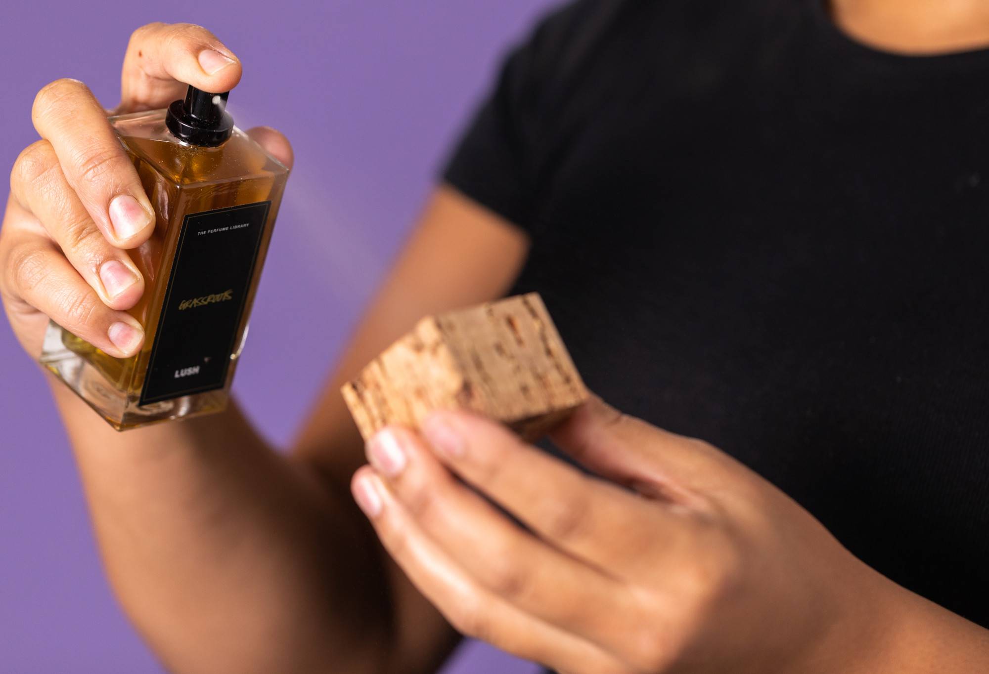 Someone spritzes a square cork lid with the black label perfume Grassroots, against a purple background.