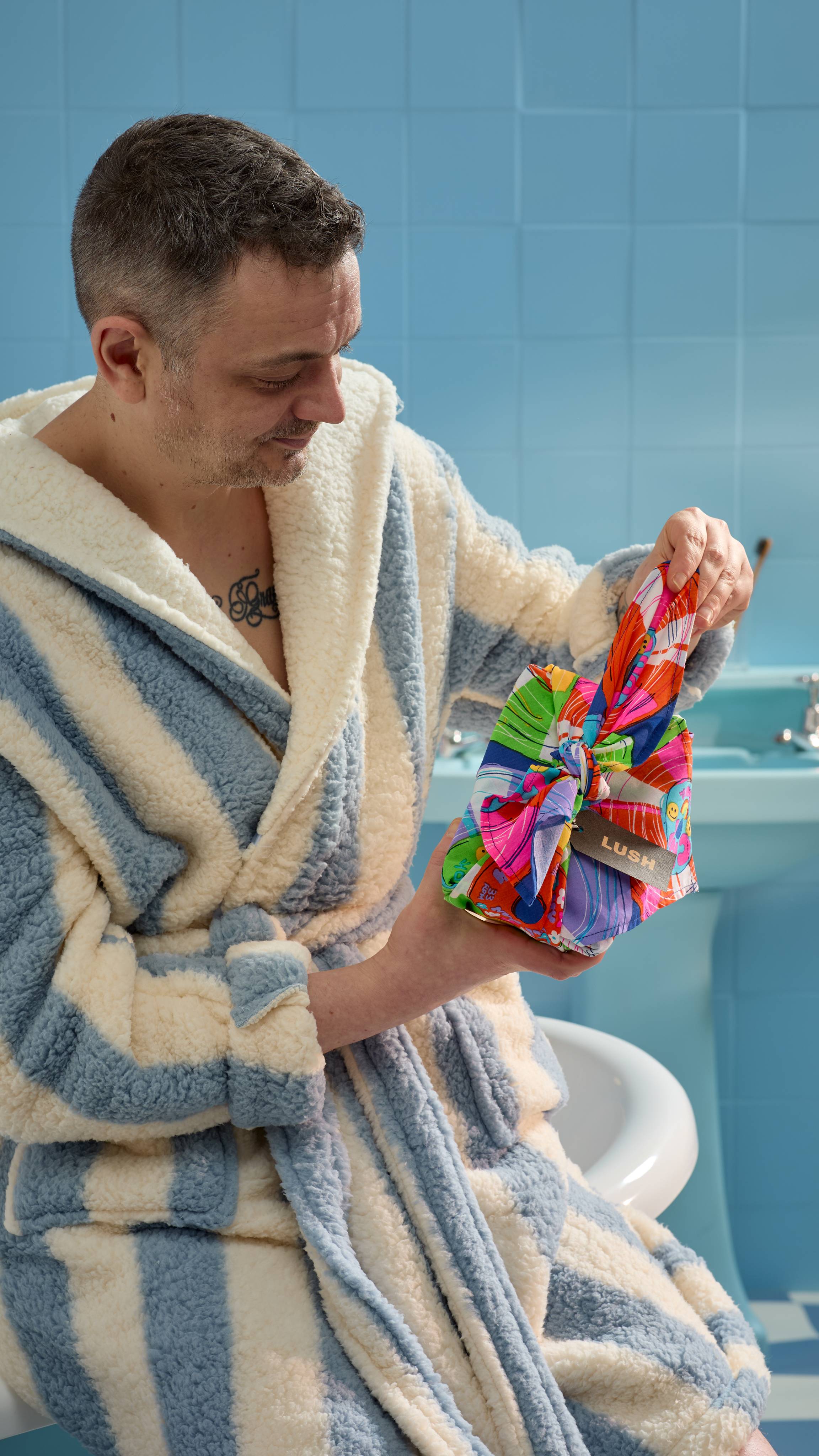 The model is sitting on the edge of the tub in a blue and white striped dressing gown as they hold the Greatest Hits knot wrap which has been wrapped neatly around some bath gifts. 