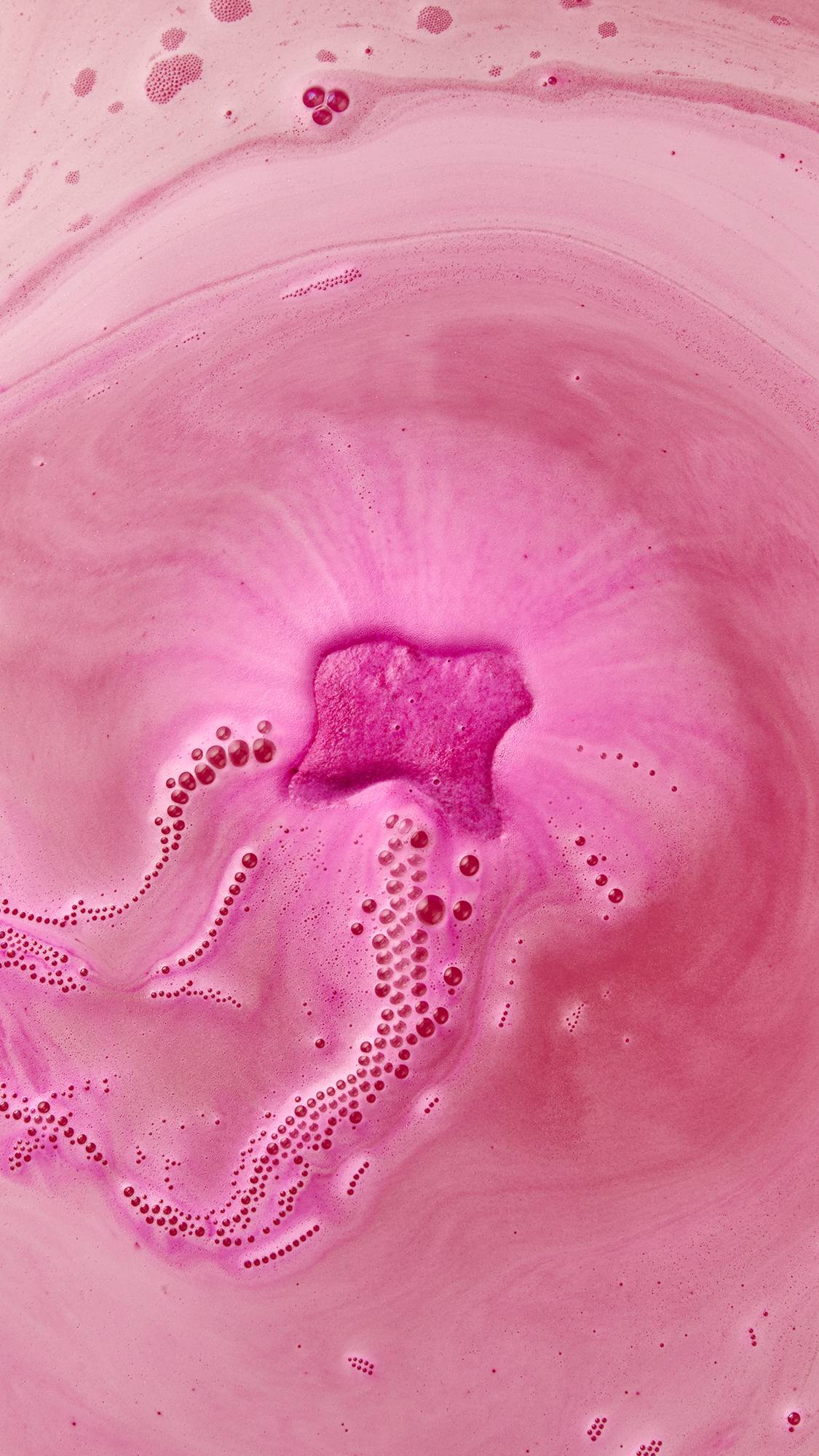 An angled, aerial shot of the Lush Groovy Snow Fairy dissolving on the surface of pink, rippled water. There is a thick, pink, velvety foam coming from the bath bomb and stretching across the surface of the water with a string of bubbles. 