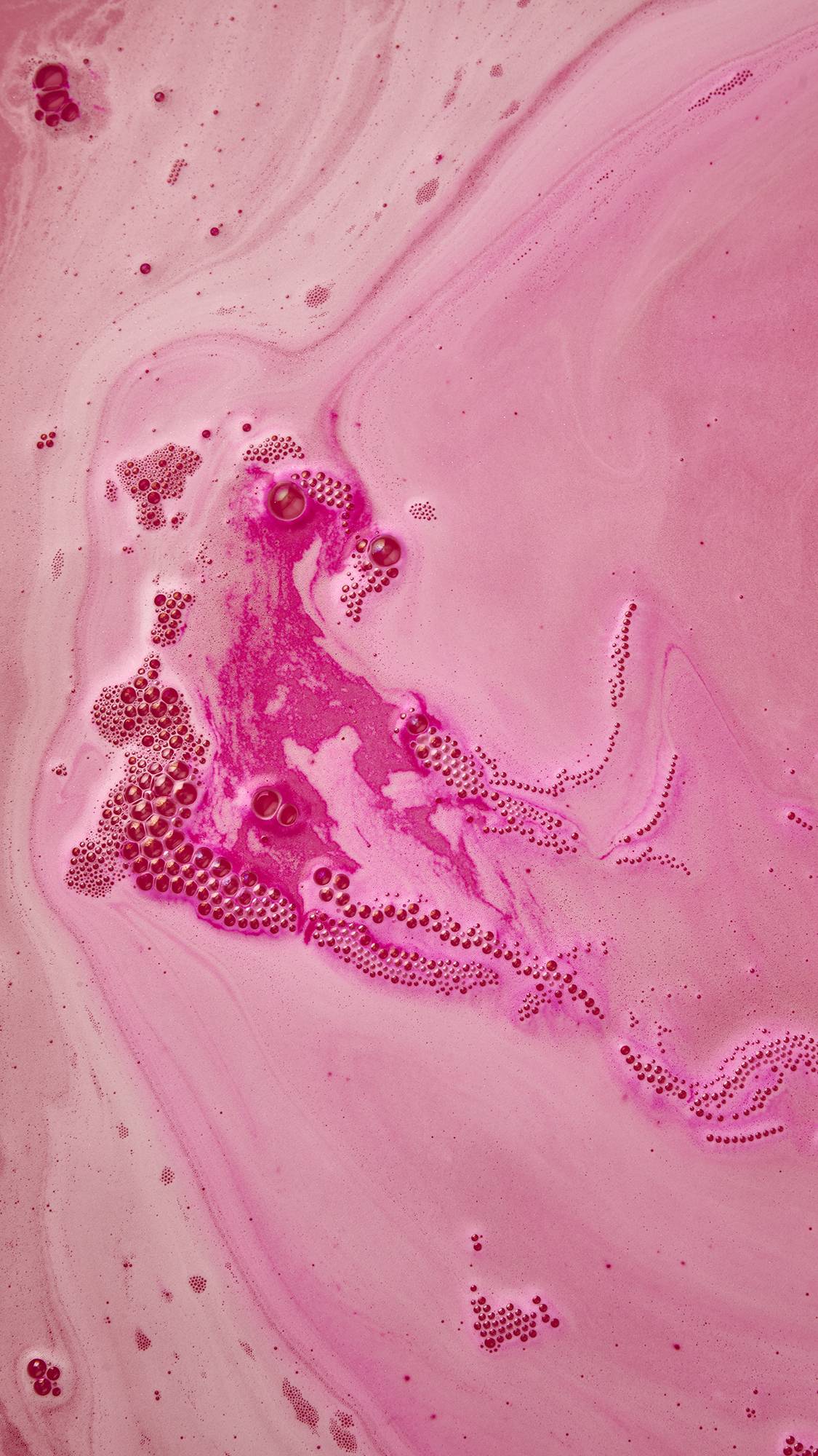 An angled, aerial shot of the Lush Groovy Snow Fairy almost fully dissolved on the surface of pink, rippled water. There is a thick, deep-pink, velvety foam coming from the centre, stretching across the surface of the water with a string of bubbles. 