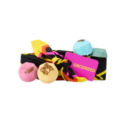 A knot-wrapped gift tagged "GROUNDED" in pink and yellow, surrounded by colourful geometric bath bombs in pink, yellow, blue, and green