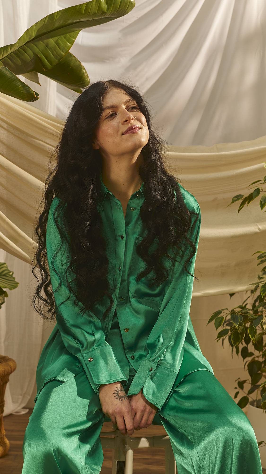 The image shows the model is long, emerald-green satin pyjamas. They are sat on a stool with a plant behind them as their long, dark, wavy hair drapes over their shoulders. 