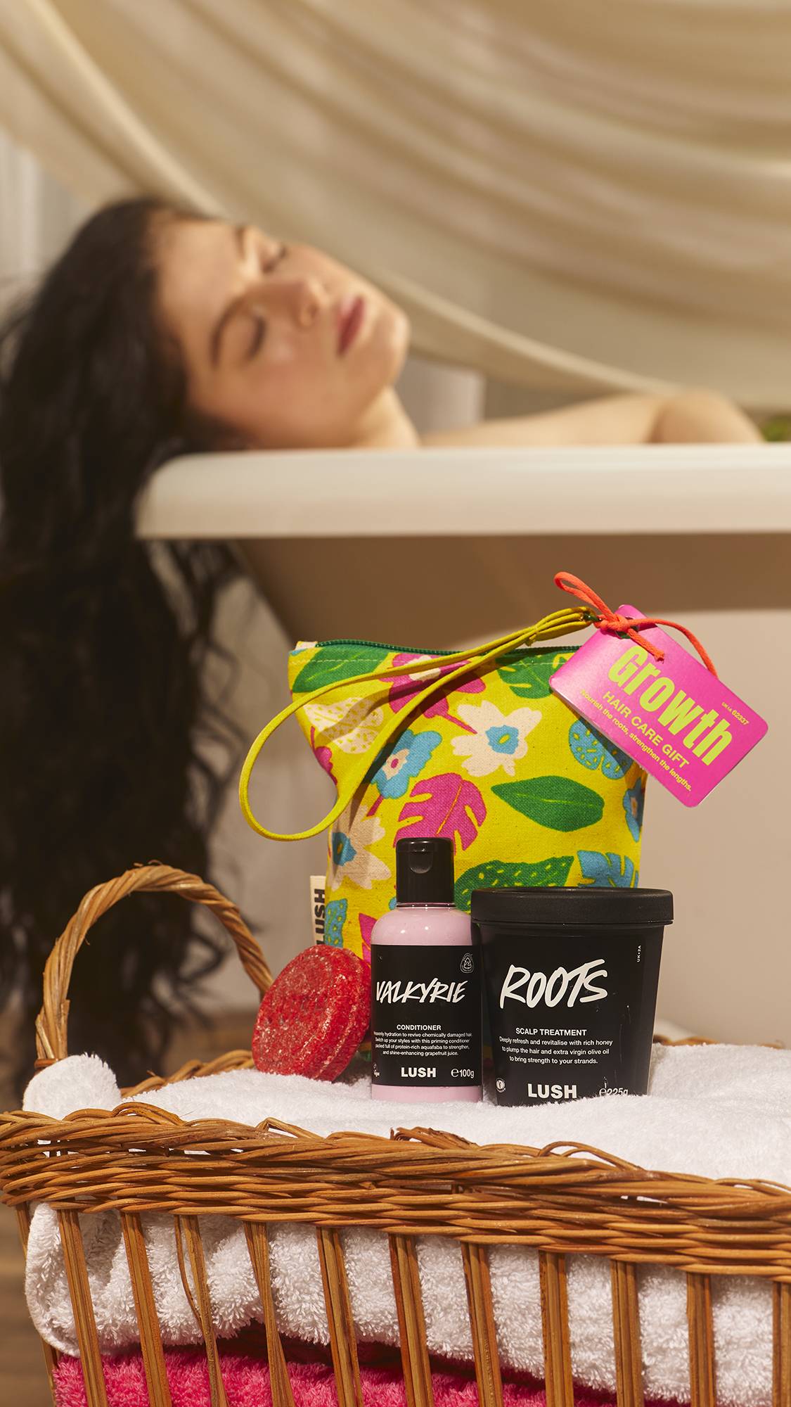 The model is laid in the bathtub relaxing in the blurred background as the foreground focuses on the haircare gift pouch and the included contents sat on a pile of folded towels. 
