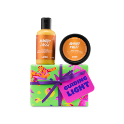 A Lush 'Guiding Light' gift set containing a bottle of Mango Lassi shower gel and a black tub of Mango Kulfi body lotion, set above a lime green wrapped gift box tied with a purple ribbon