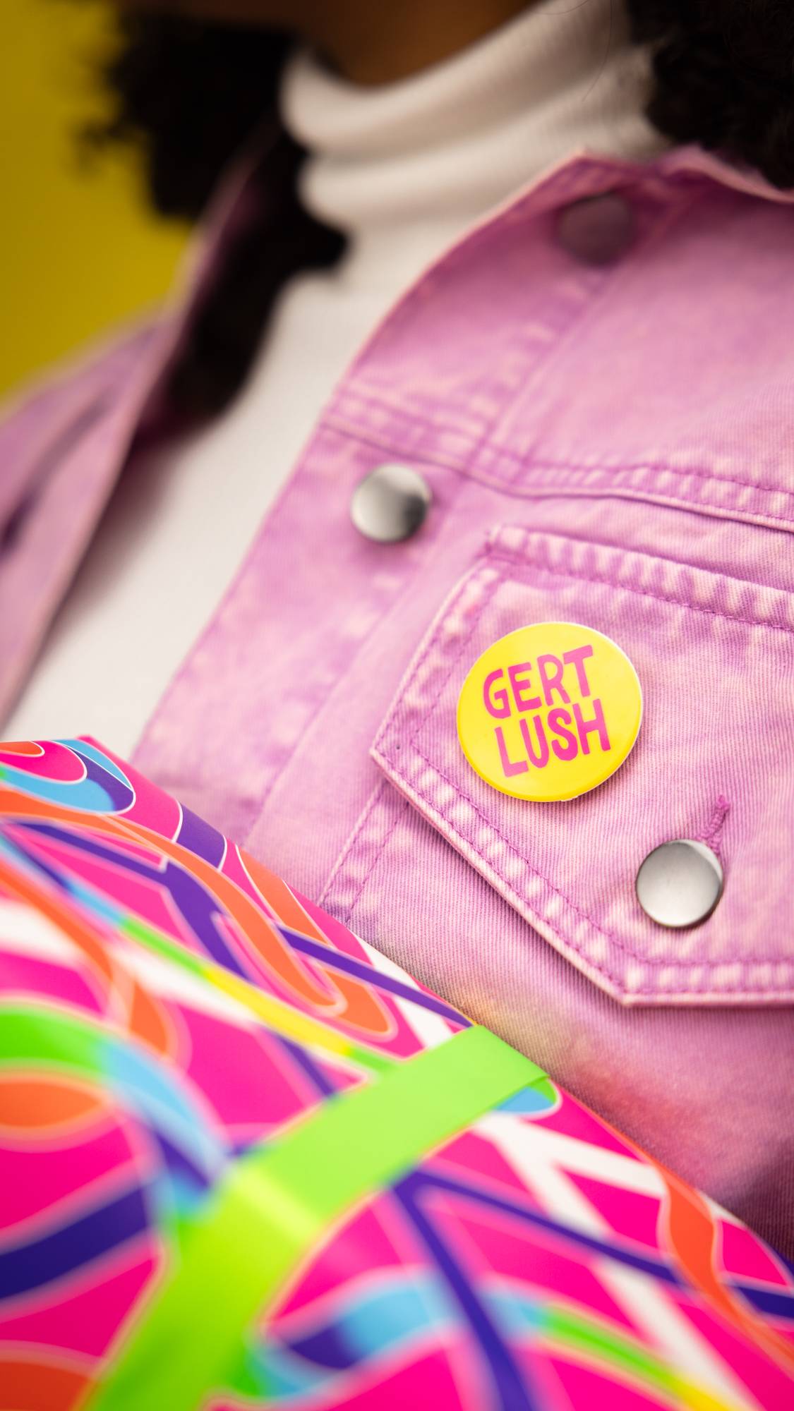Image shows model wearing a fuchsia-coloured, denim jacket. A yellow, pinned badge shows the words "Gert Lush" written in pink.