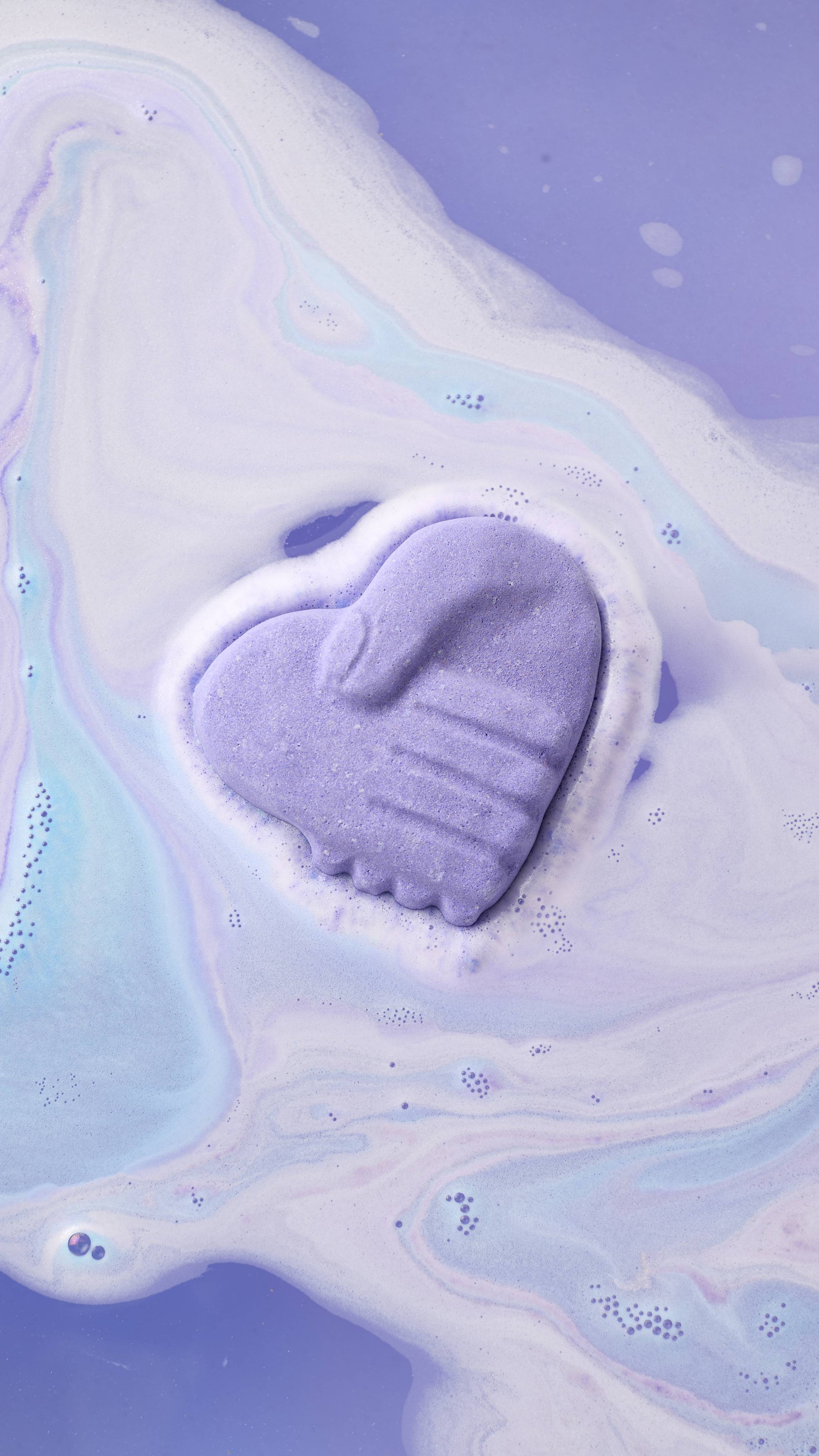 A purple, heart-shaped bath bomb with a molded design of two hands shaking floats in a bath. It is surrounded by swirling patterns of light blue and lavender foam as it dissolves into the deep purple water.