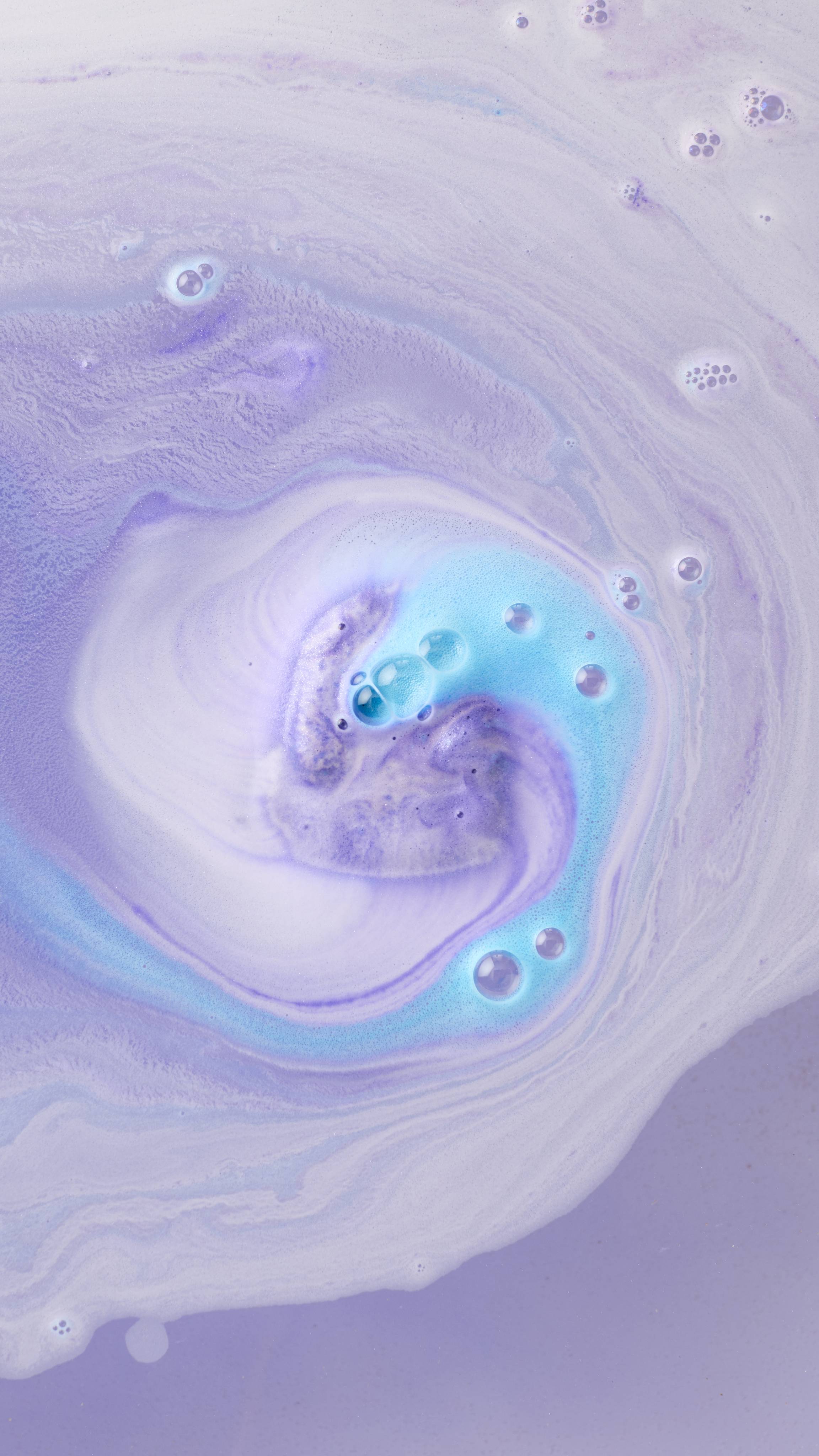 A top-down view of bathwater after a bath bomb has dissolved, featuring a swirling galaxy of pastel purple, periwinkle, and white foam. Vibrant turquoise bubbles cluster in the center of the whirlpool, creating a dreamy, marbled effect.