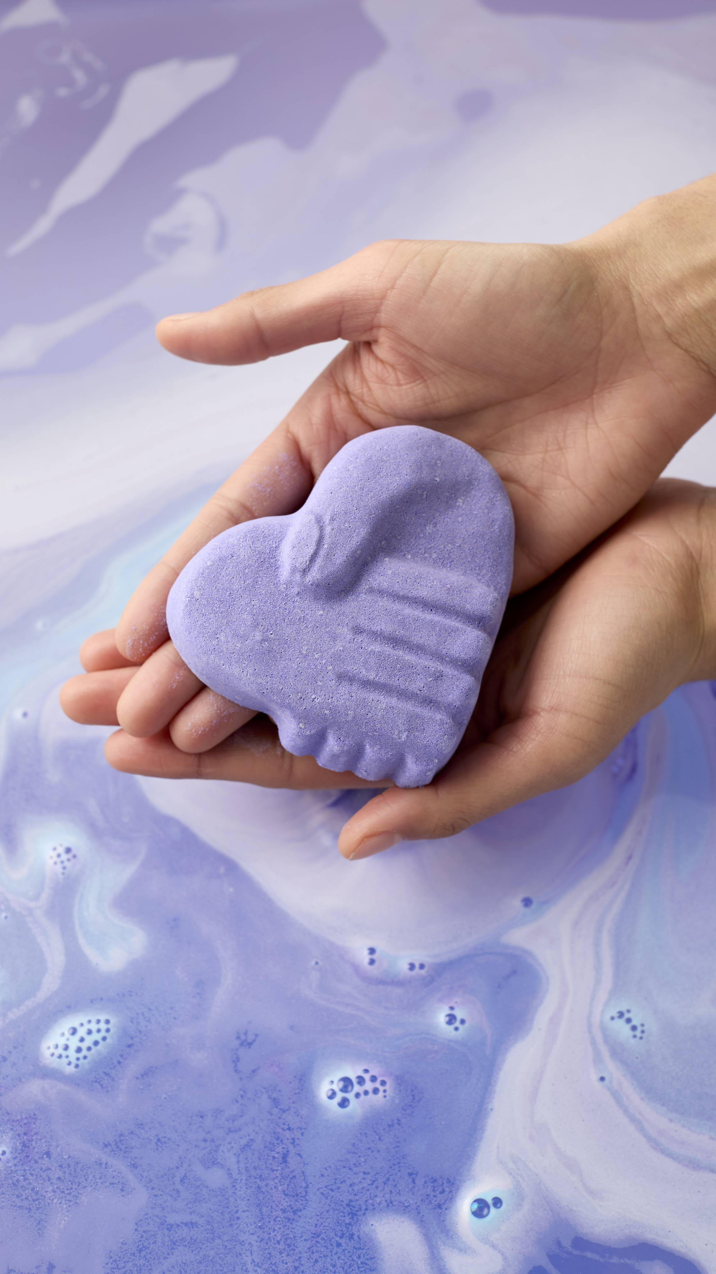 A pair of hands holds a purple, heart-shaped bath bomb featuring a handshake design. It is held over a bath of swirling lavender water with patterns of pale blue and white foam.