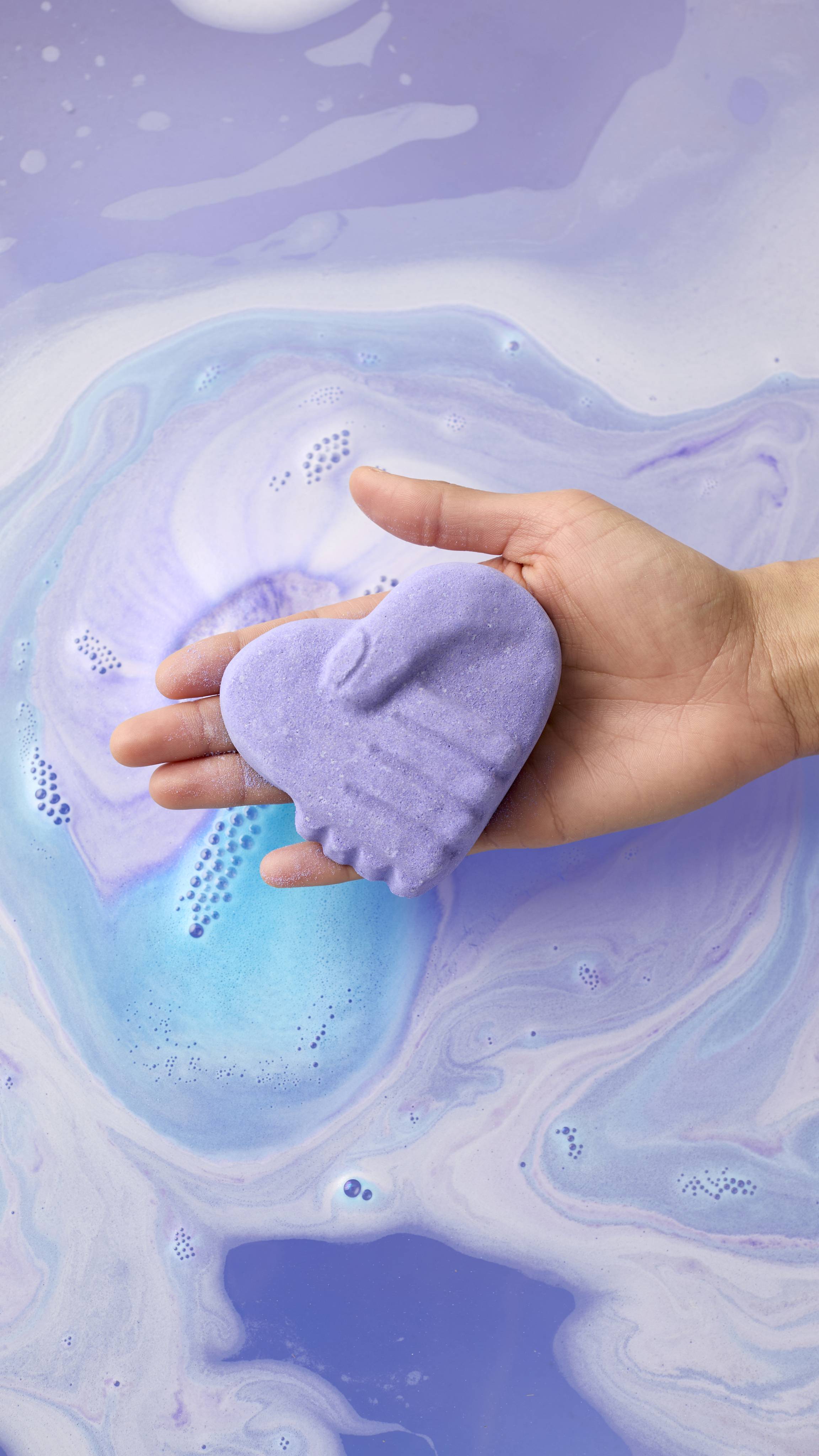 A hand holds a purple, heart-shaped bath bomb with a molded image of two hands shaking. It is held over swirling purple and blue bathwater as it begins to dissolve, creating a marble-like pattern of colors.