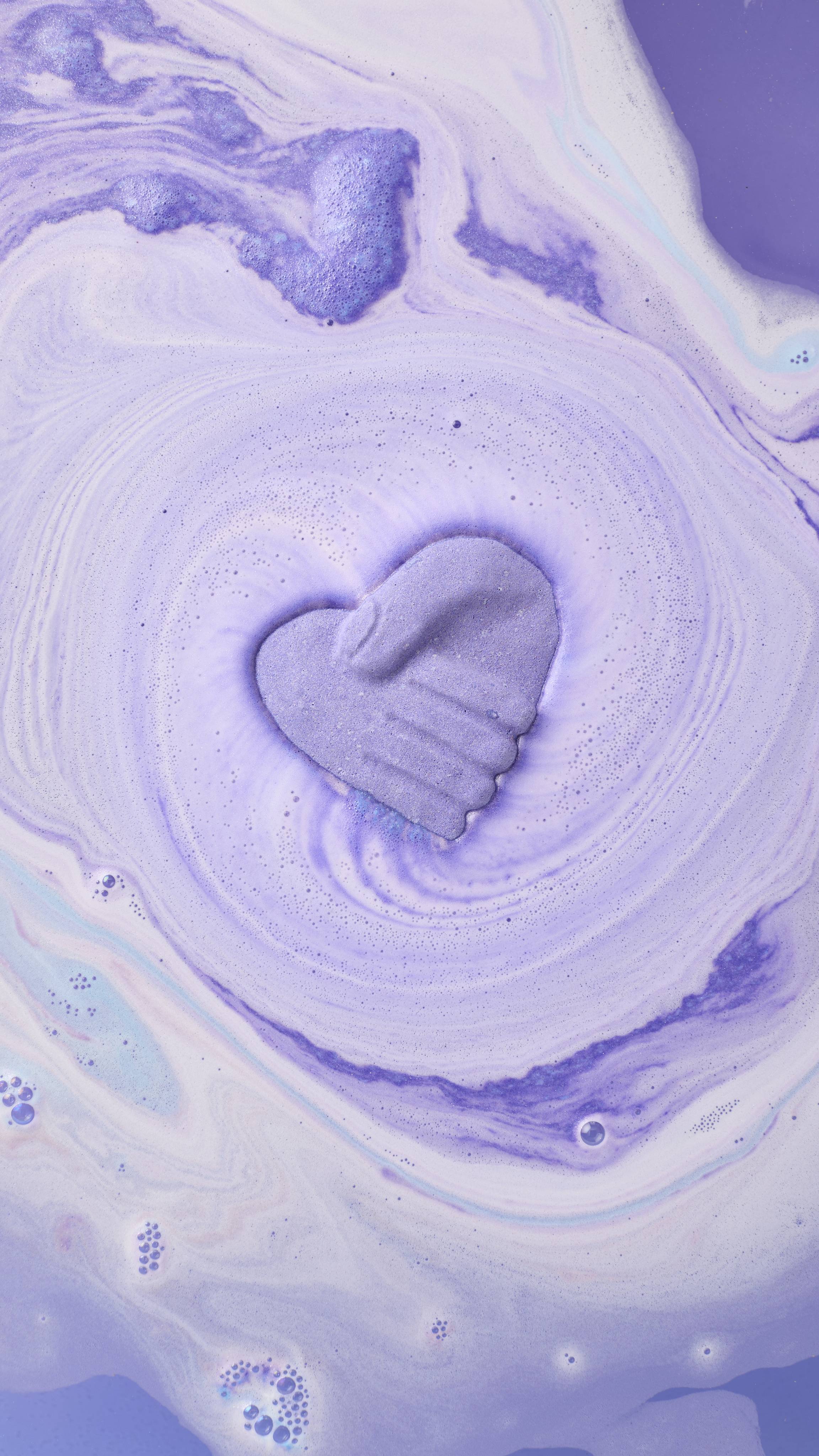A purple heart-shaped bath bomb with a handshake design spins in a bath, creating a whirlpool of lavender and periwinkle foam. The surrounding water is a deep, tranquil purple with delicate white swirls.