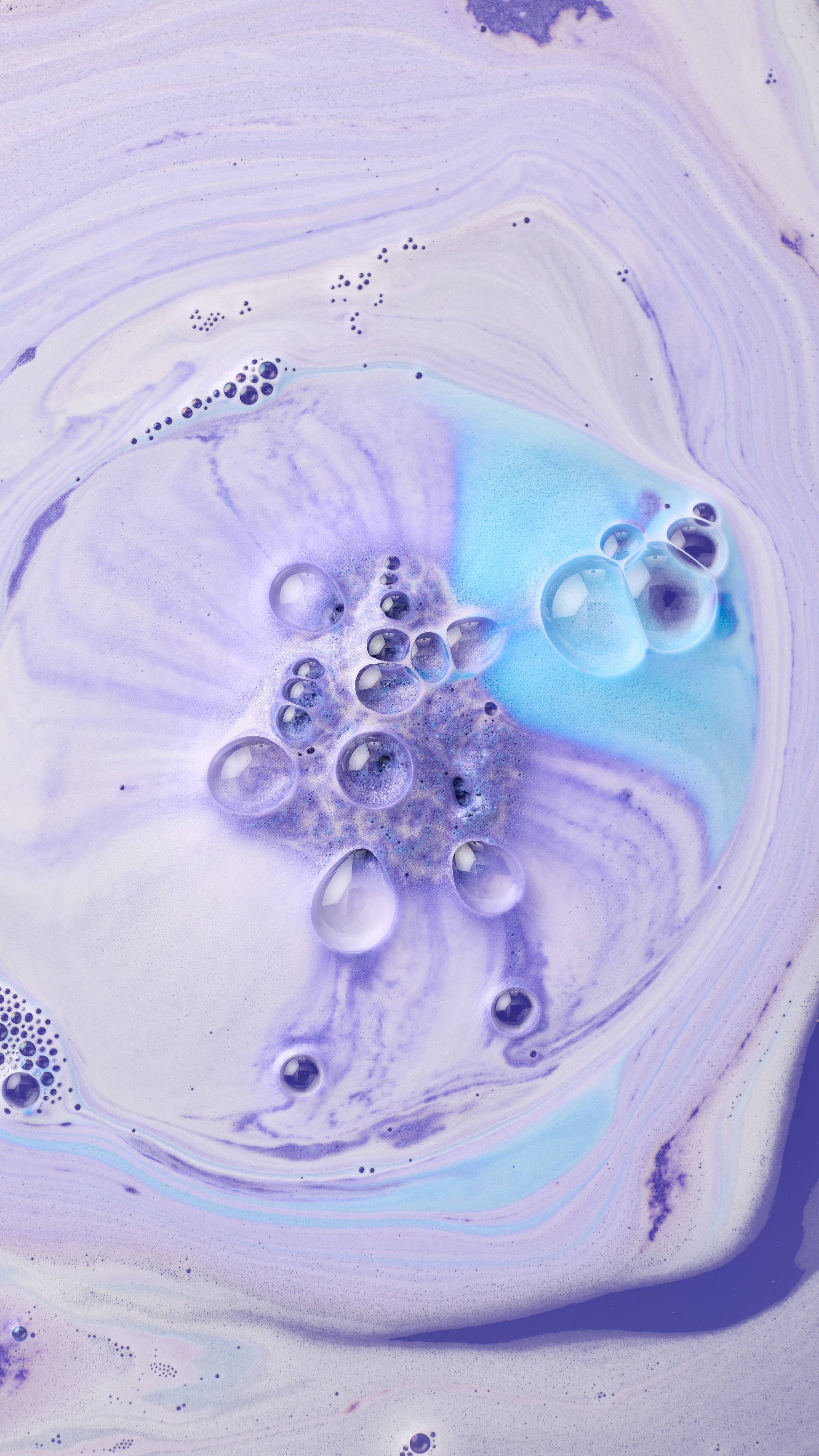 A top-down view of bathwater featuring a mesmerizing swirl of pastel purple and periwinkle foam. Large, clear bubbles and bright turquoise foam are concentrated in the center, creating a vibrant, marble-like texture across the surface.