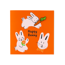 Happy Bunny. An orange greeting card features three hand-drawn pink rabbits and the text "Happy Bunny". One rabbit carries a carrot, another holds an orange flower, and a third wears a polka-dot bow tie. The carrot is a removable pin badge.