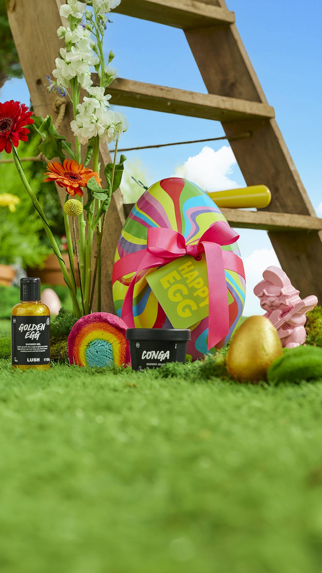 The image shows the bottom of the wooden ladder surrounded by grass, moss and flowers as the Happy Egg gift box and products are sat on the floor. 