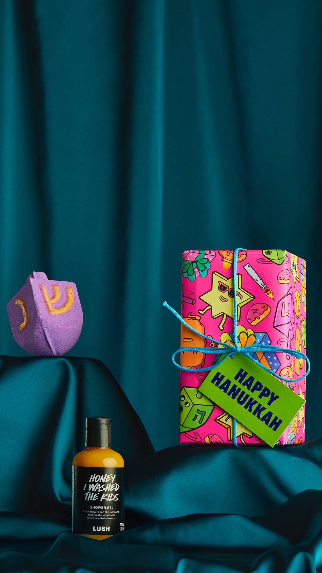 The Happy Hanukkah gift box, themed bath bomb and shower gel are sat at differing heights on top of a satin teal cloth. 