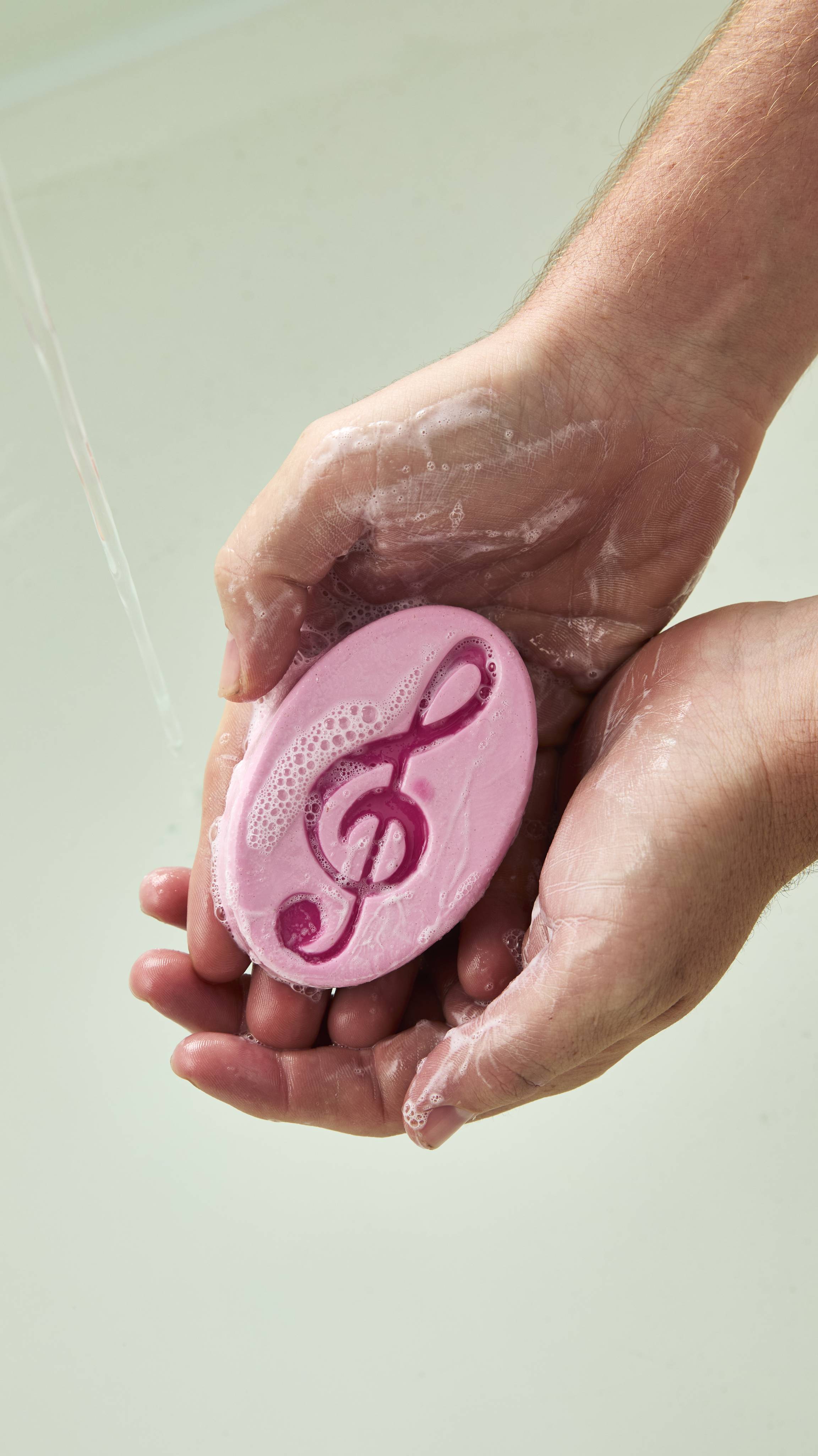 Hands lather an oval pink Lush "Harmony" soap bar with a deep pink treble clef symbol under a stream of running water, creating suds over their skin.