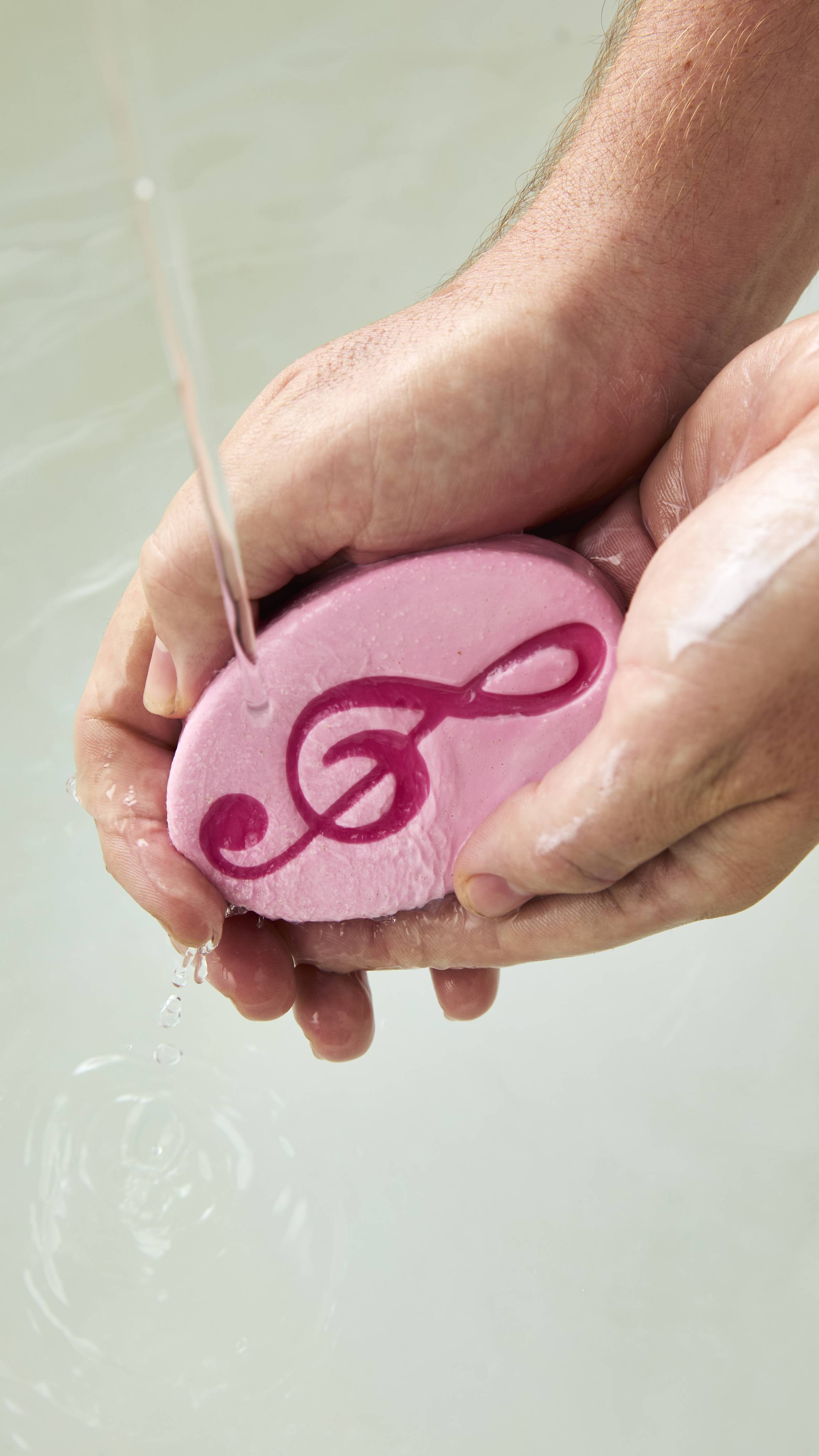 Hands lather an oval pink Lush "Harmony" soap bar with a deep pink treble clef symbol under a stream of running water.