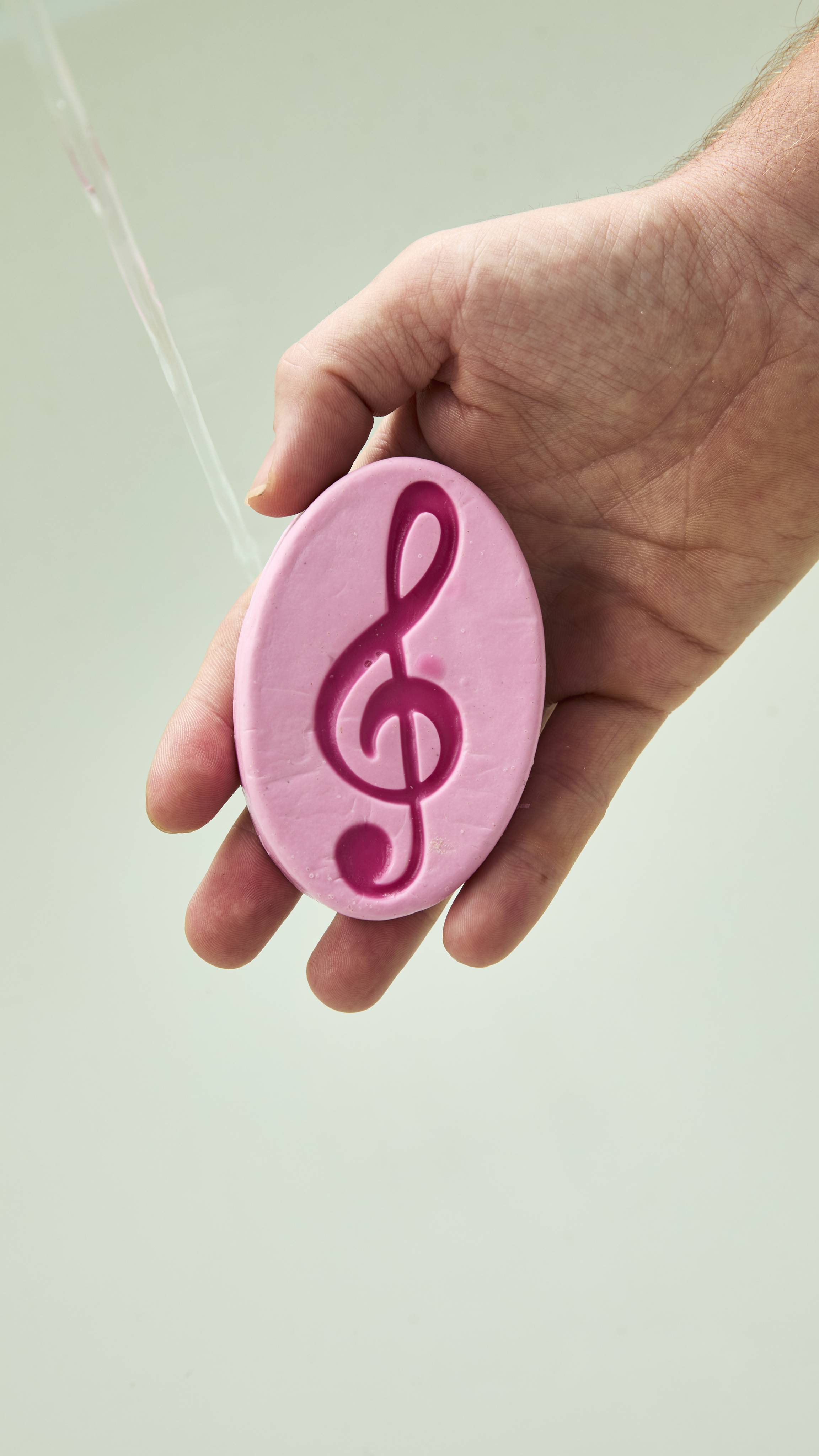 An open hand holds an oval pink Lush "Harmony" soap bar featuring a deep pink embossed treble clef symbol, held beneath running water in front of a neutral backdrop.