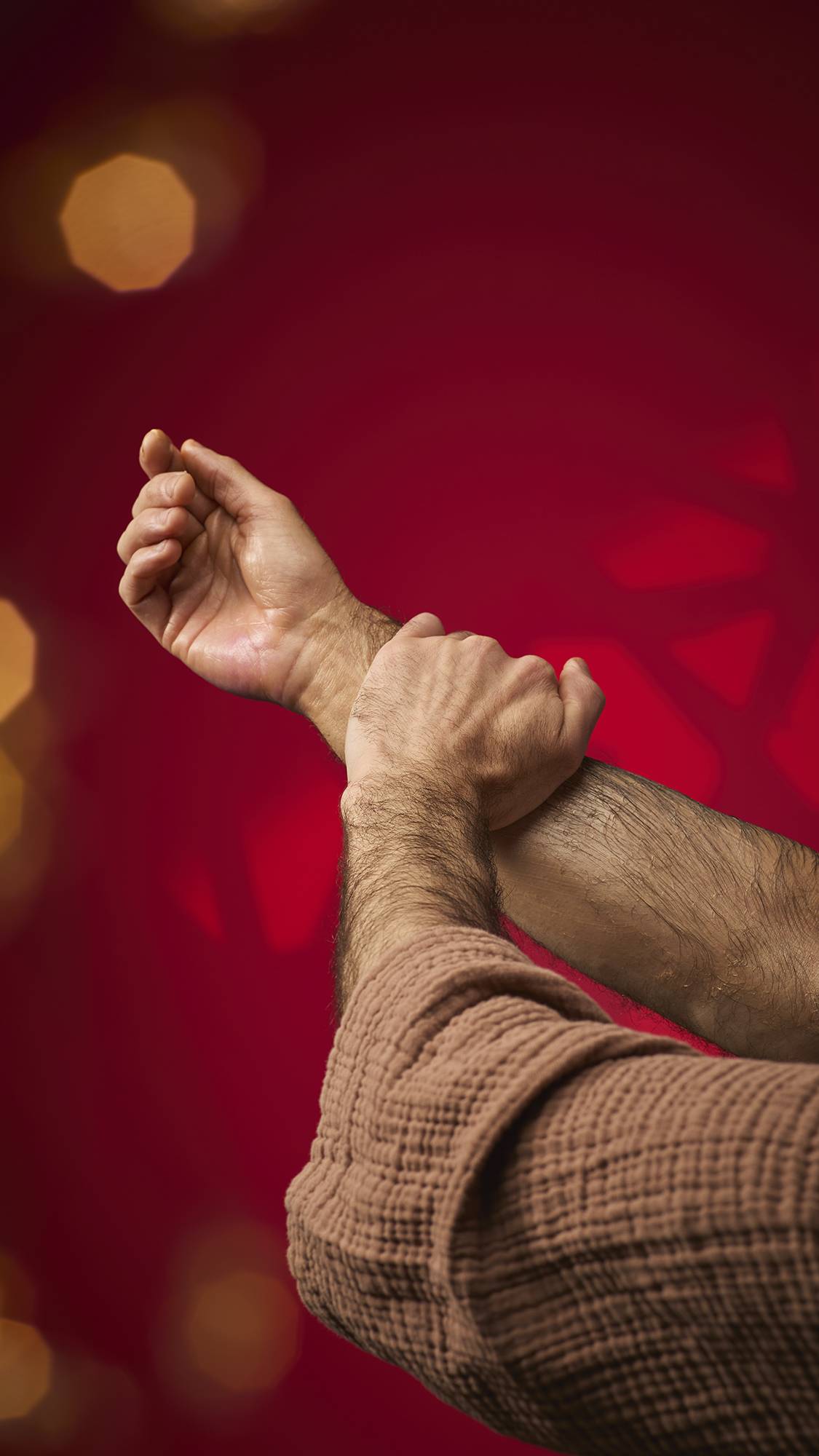A person massages a shimmering, golden oil from the Hayl massage bar into their forearm. Their skin has a radiant, glowing finish, and they are wearing a brown textured robe against a deep red background with soft golden bokeh lights.