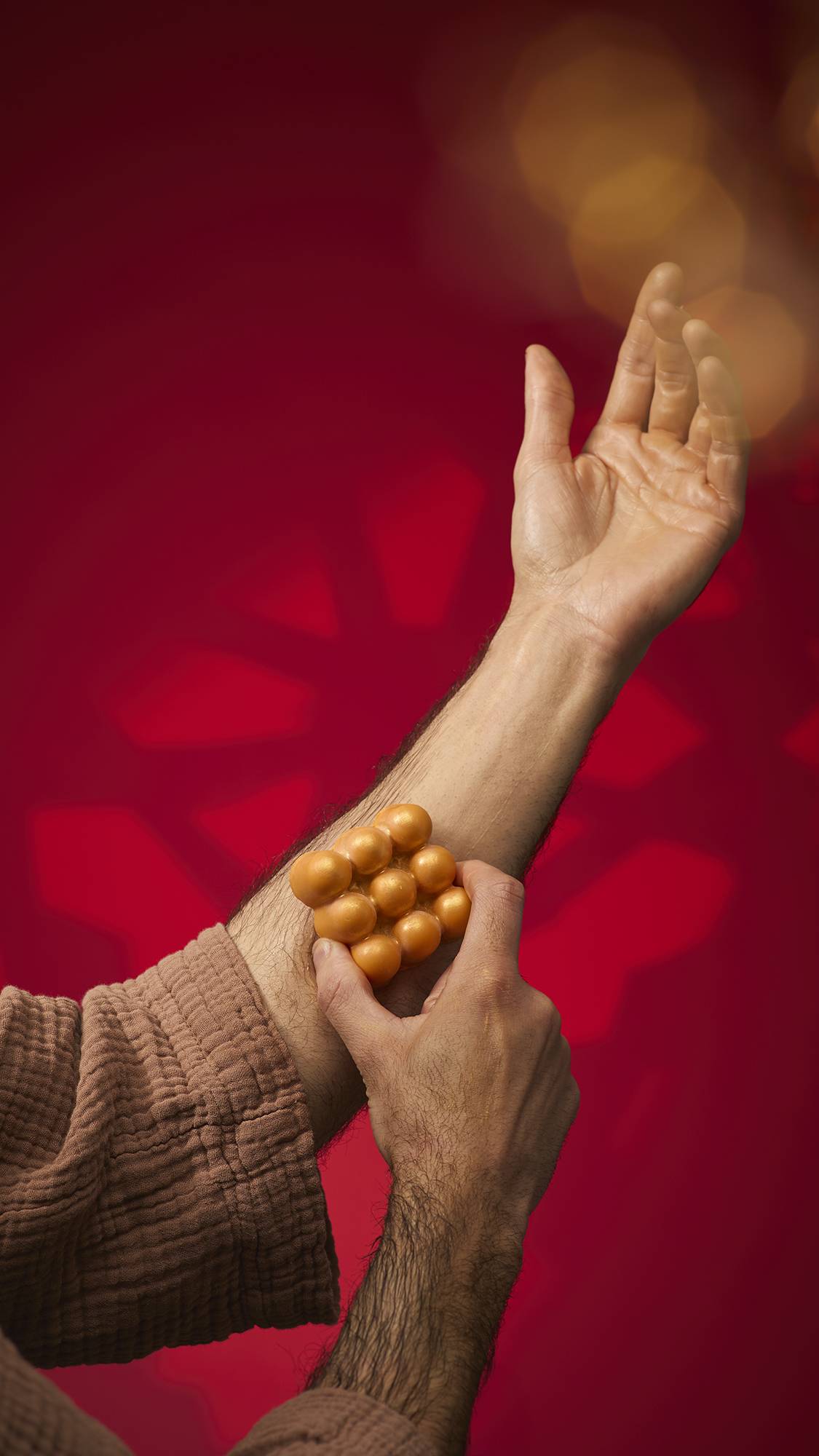 A person uses the golden, shimmering Hayl massage bar by pressing its rounded, bulbous surface against their forearm. They are wearing a brown textured robe against a deep red background with soft gold bokeh lights.