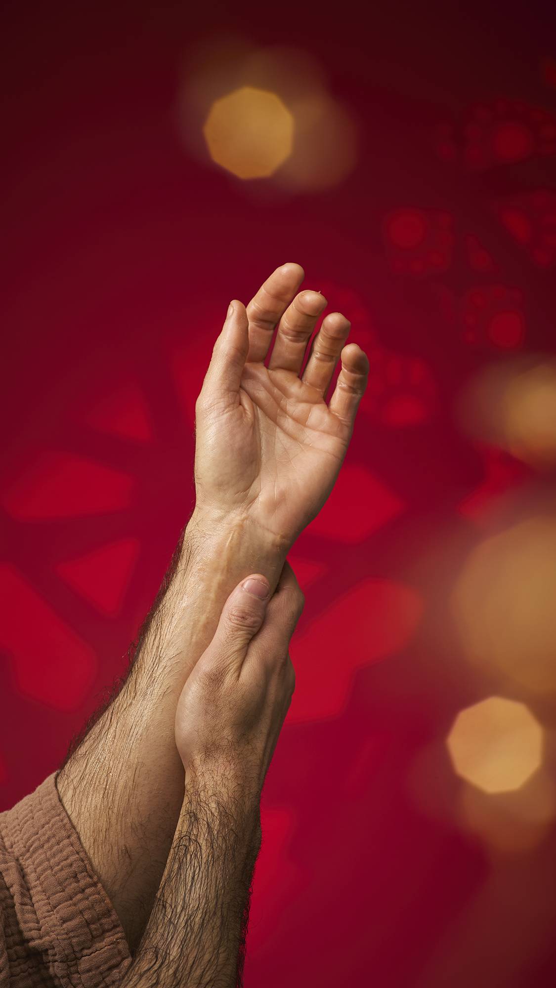 Hands massage shimmering oil from the Hayl massage bar into an arm, leaving a radiant glow. The person wears a brown textured robe against a deep red background with golden bokeh lights.