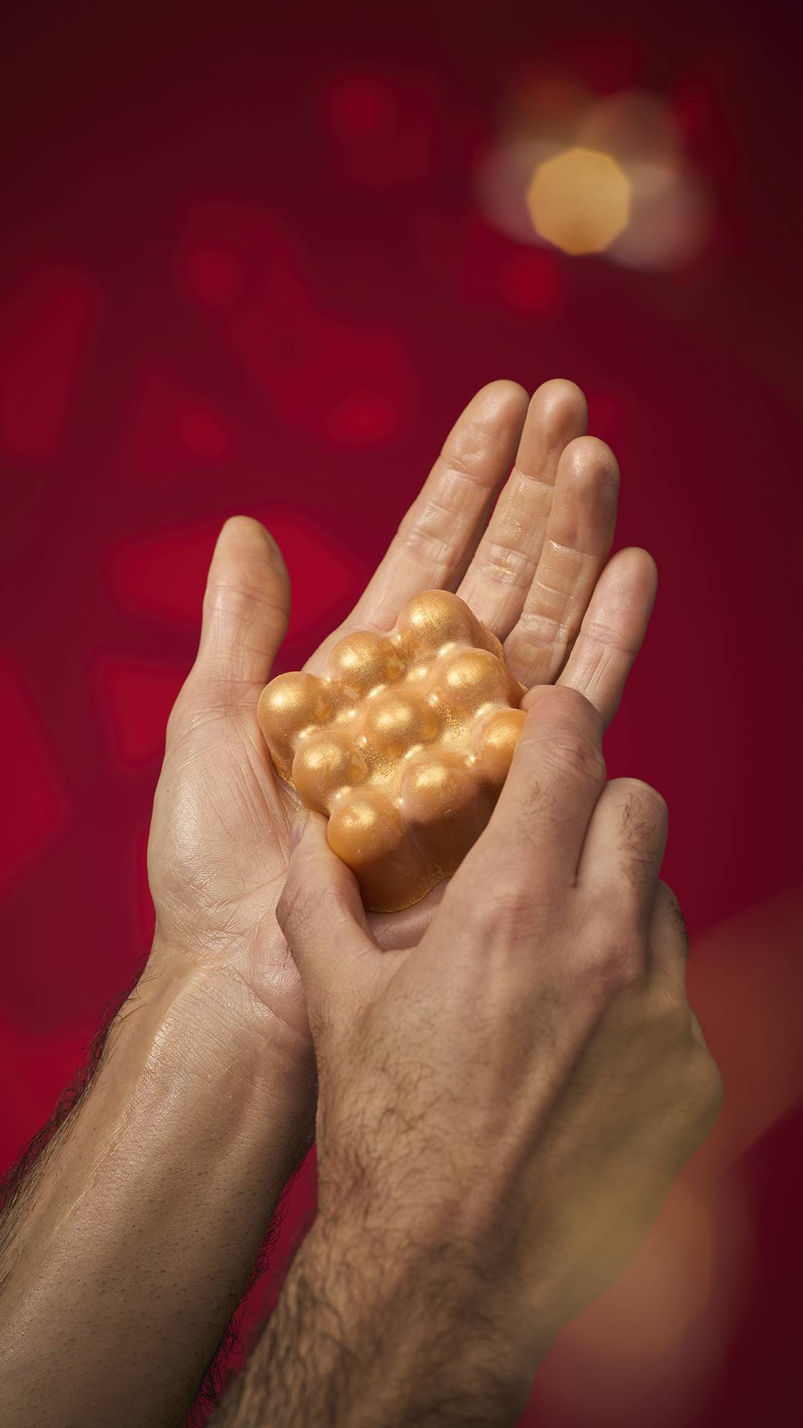 Hands hold the shimmering golden Hayl massage bar. The bar's bulbous surface leaves a radiant oil glow on the skin. Set against a deep red background with golden bokeh lights.