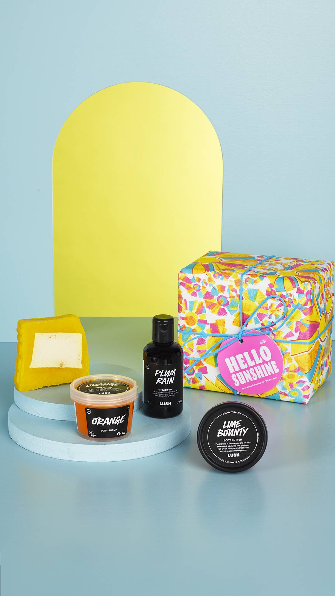 The vibrant, sun-patterned gift box and the four LUSH products sit on a pastel blue floor and background with an arched mirror in the background.