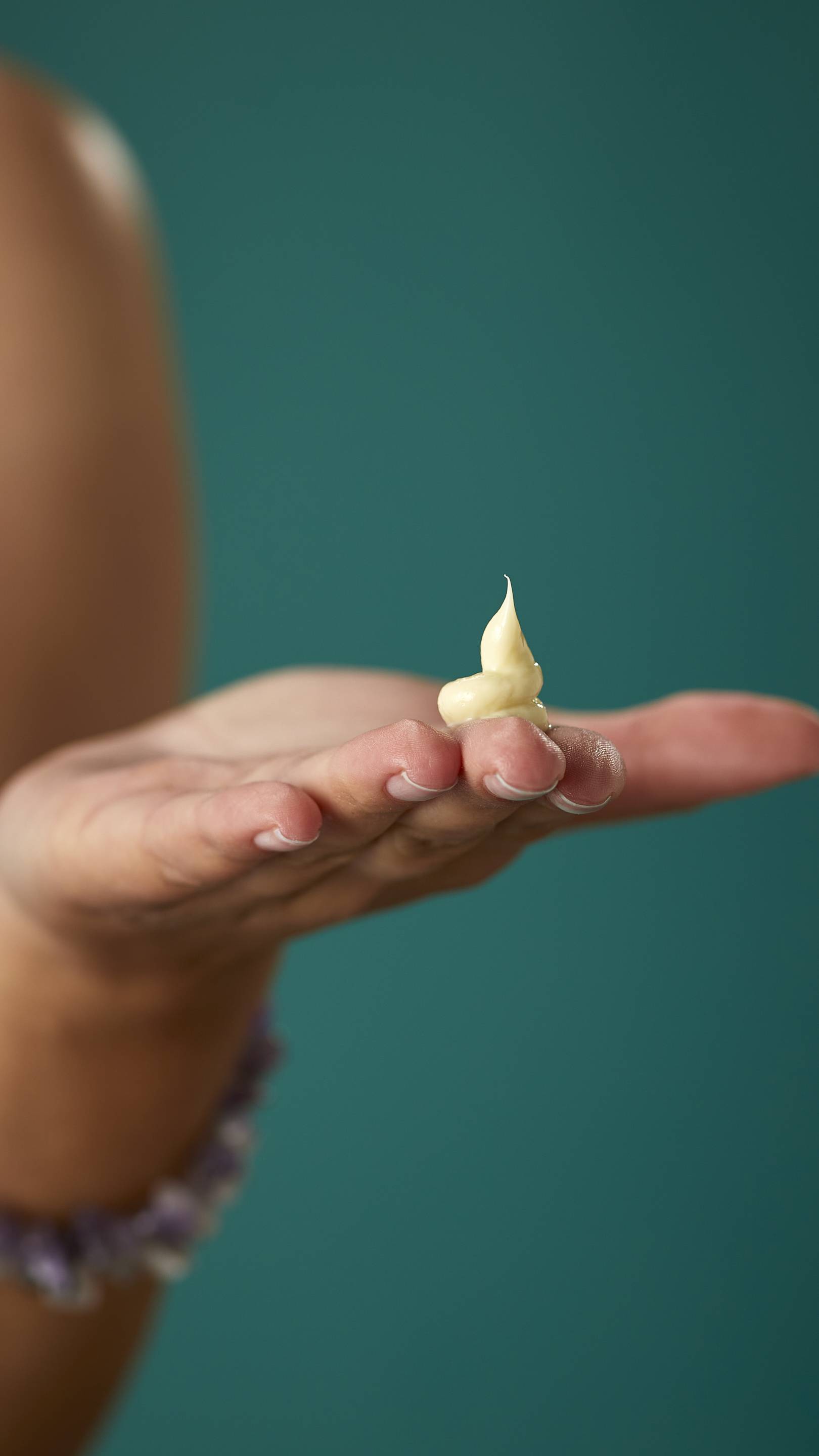 The image shows a light-toned hand with a flat palm, holding a perfectly formed dollop of pale cream product with a peak. The hand, with pale nails and a beaded bracelet, is against a dark teal background.