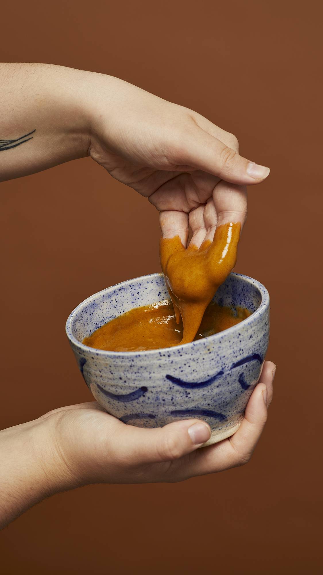 Image shows the model scooping the Hennaed hot oil treatment out of a blue ceramic bowl in front of an earthy background.